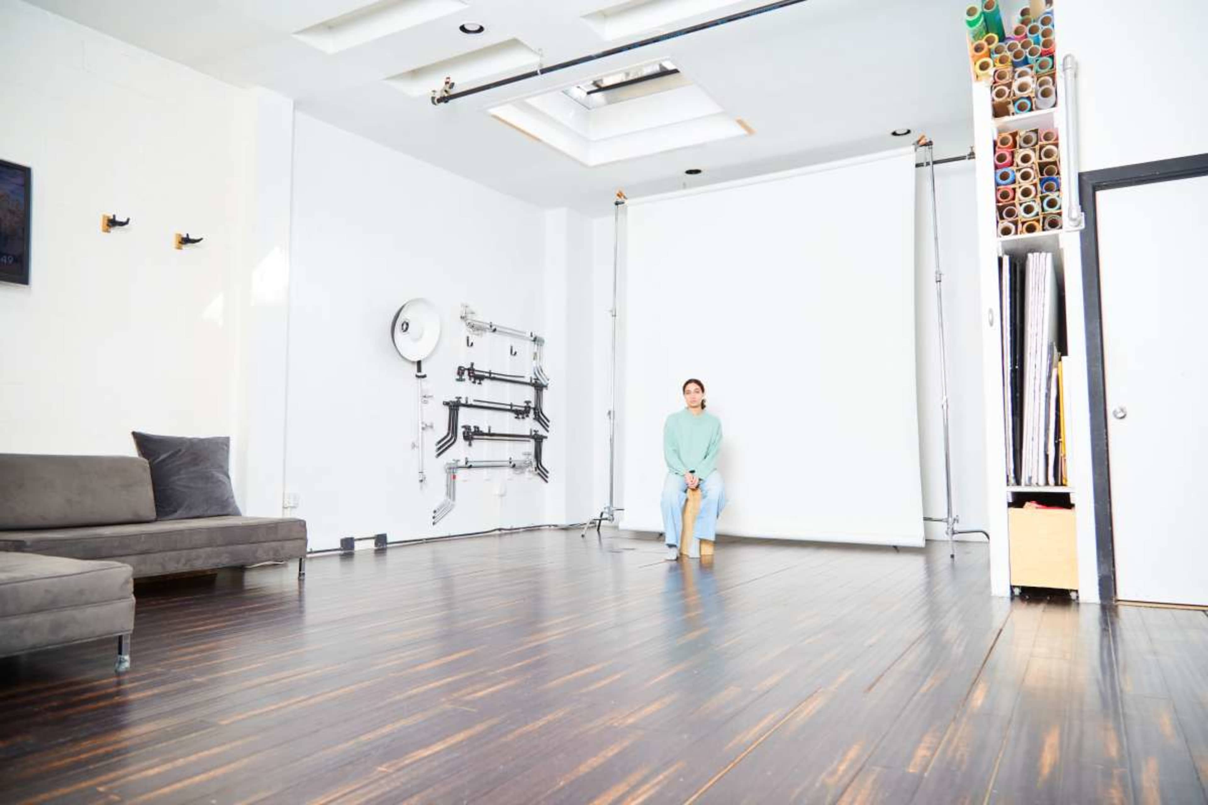 A person sits on the floor in a spacious photo studio with a white backdrop and wooden flooring.