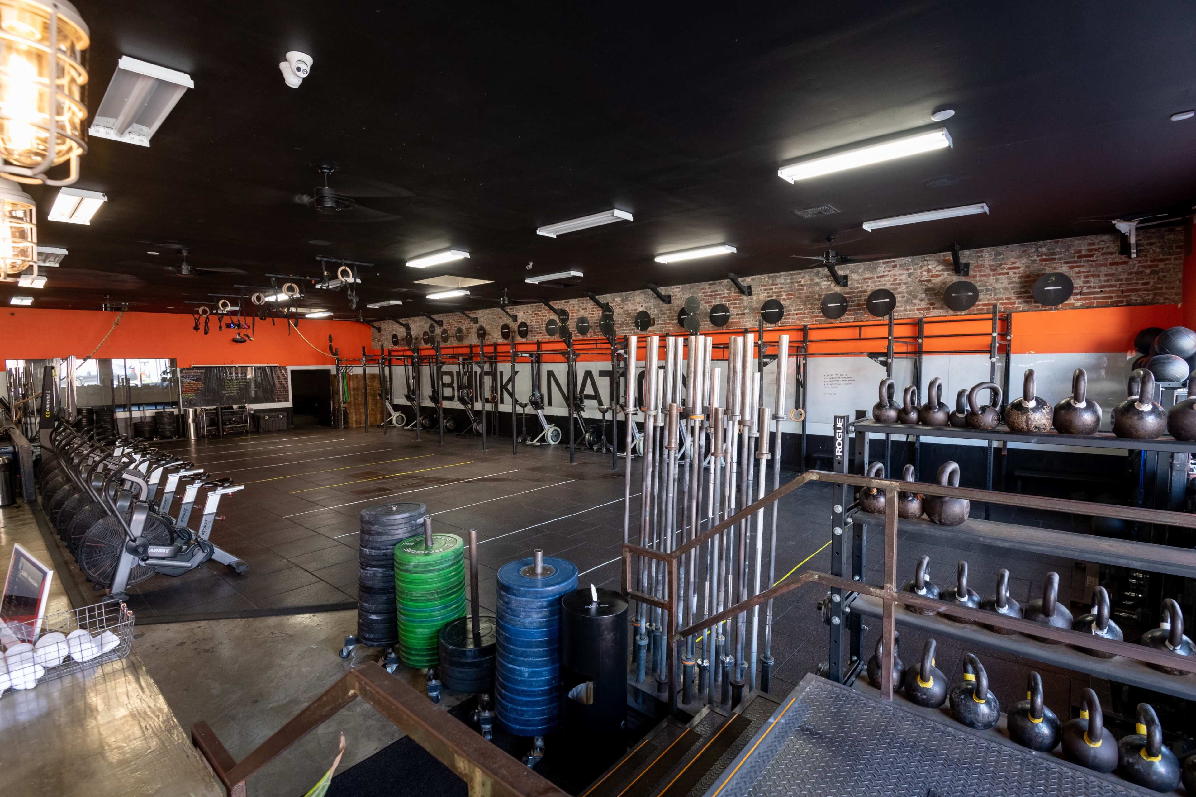 The image shows a well-equipped gym space featuring various fitness equipment, including weightlifting bars, kettlebells, and rowing machines, set against a brick wall.