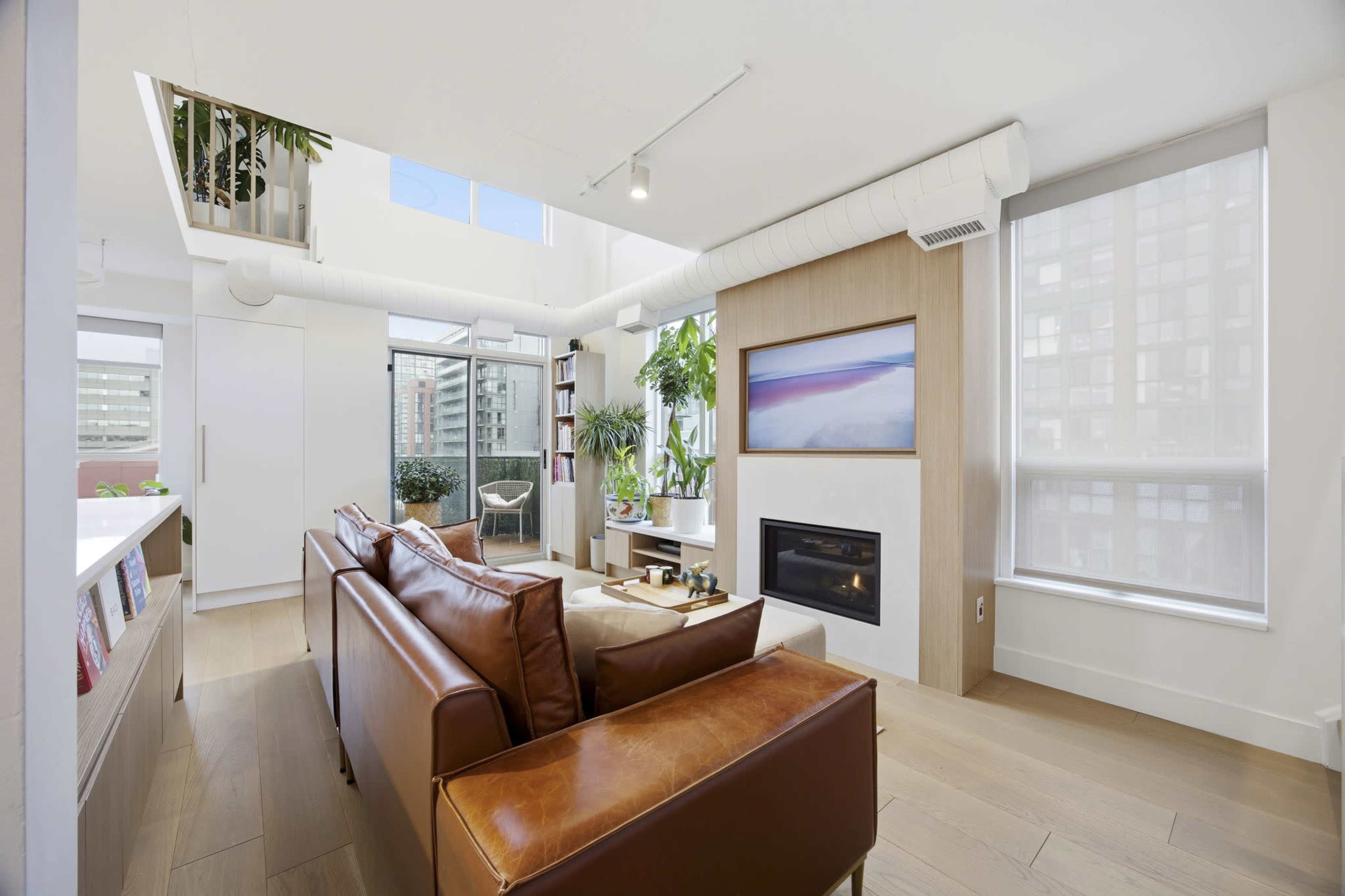 The image shows a bright, modern living room with a leather sofa, a fireplace, and large windows that provide natural light and a view of a balcony.