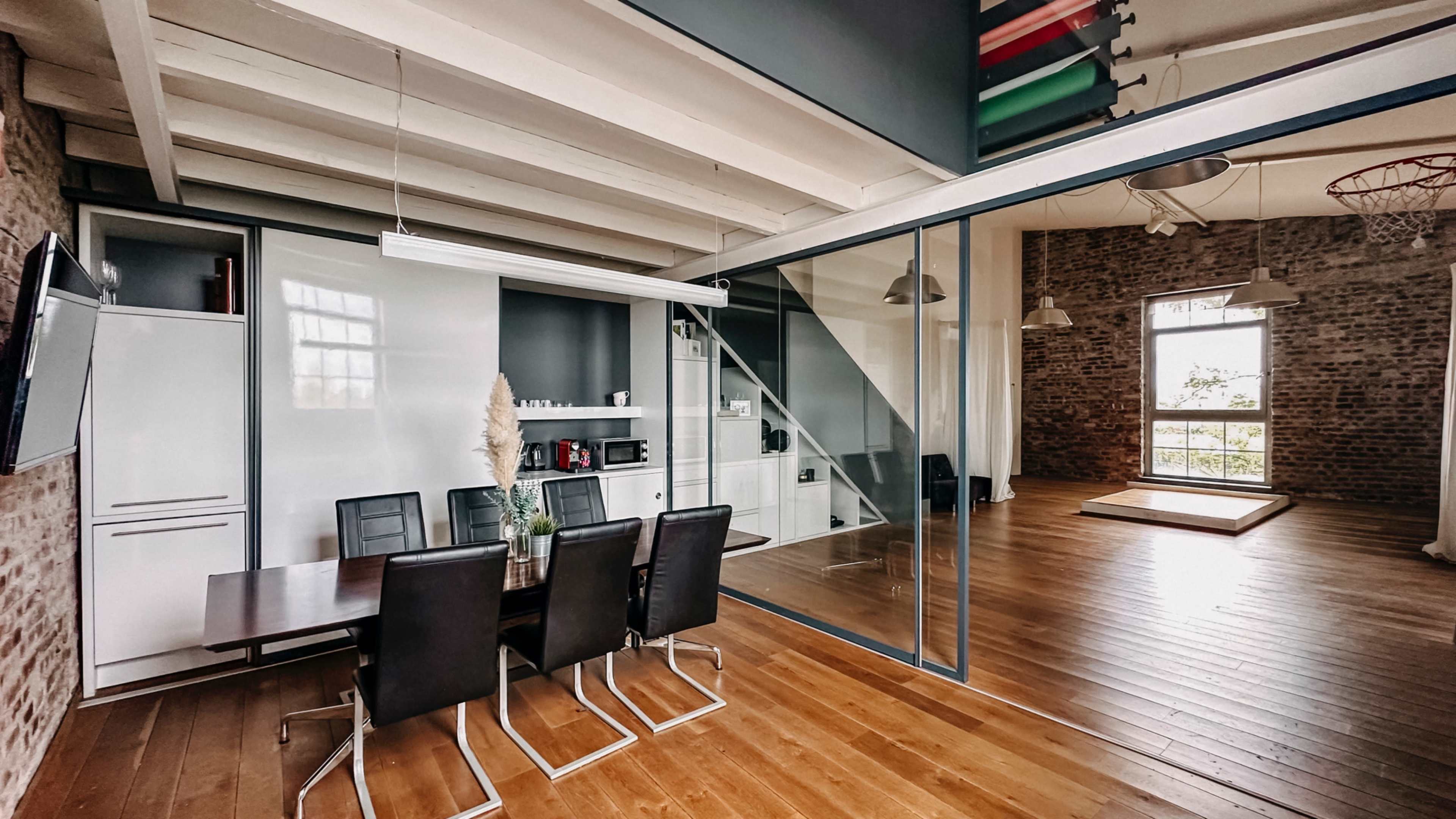 The image shows a modern office space featuring a dining table with chairs, a glass partition, and a large window with natural light.