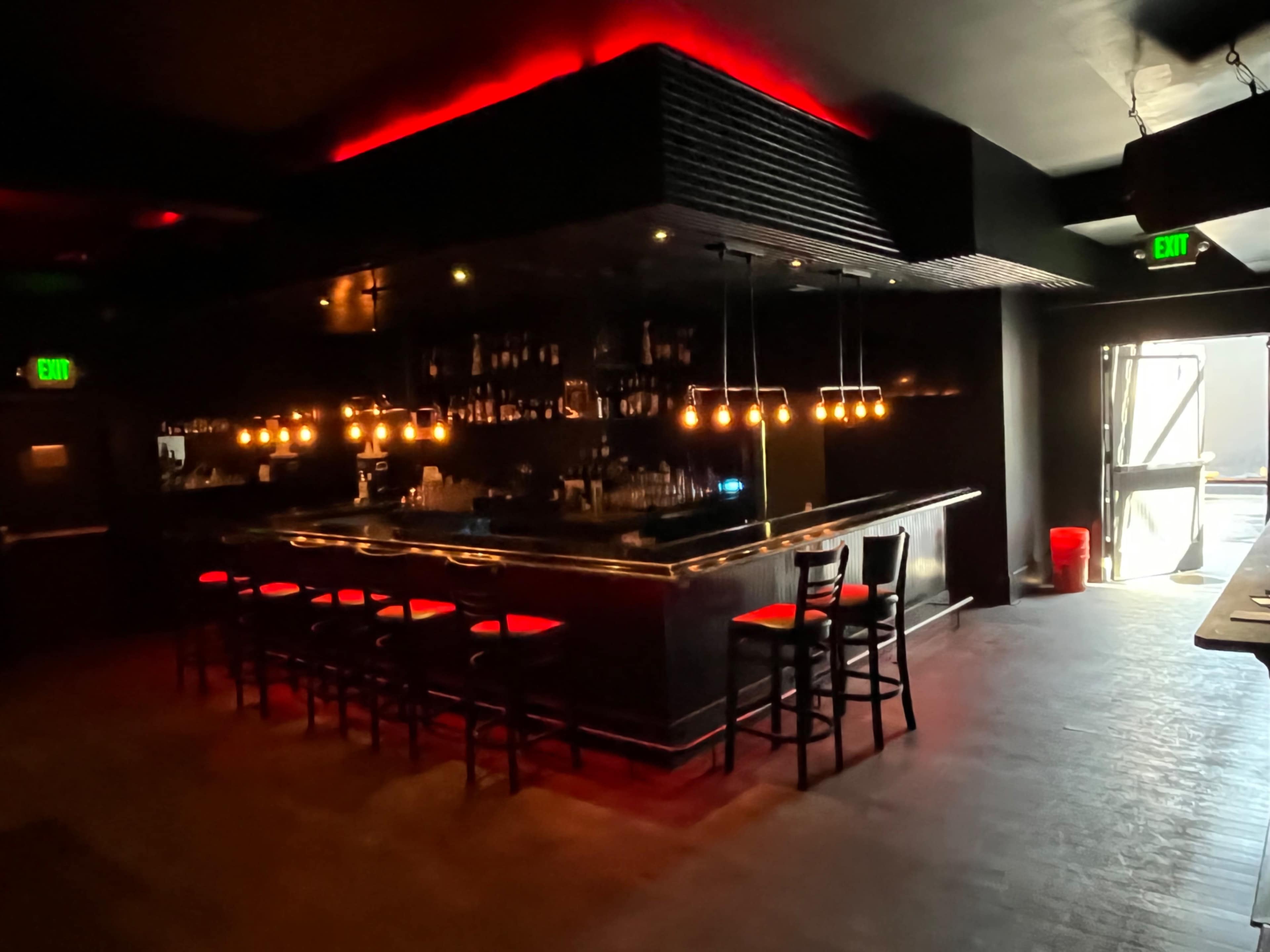 A dimly lit bar features a long counter with a row of chairs and pendant lights above, while an open door leads to an exterior area.