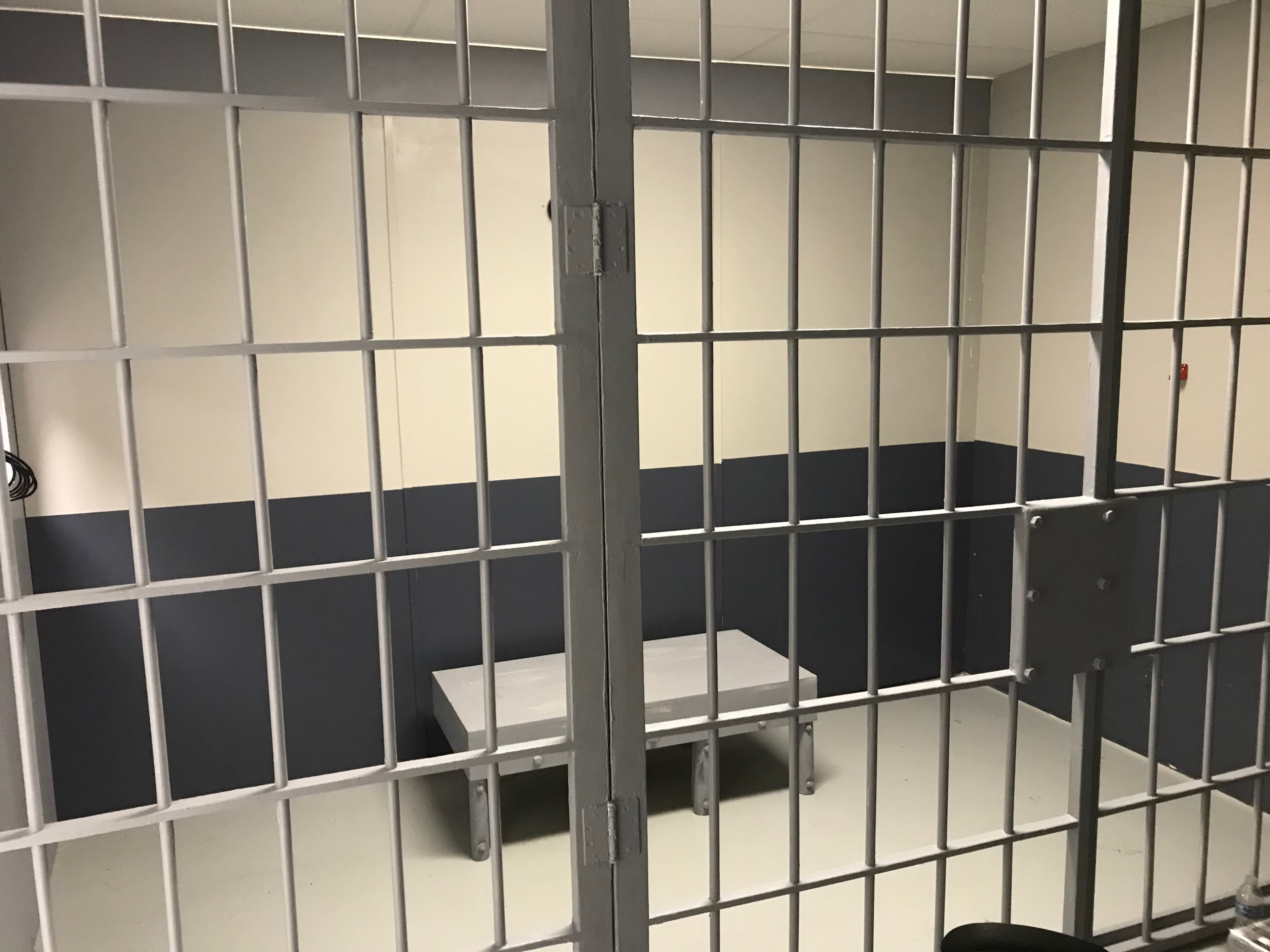 The image shows a small, empty jail cell with a gray table in the center, enclosed by metal bars.