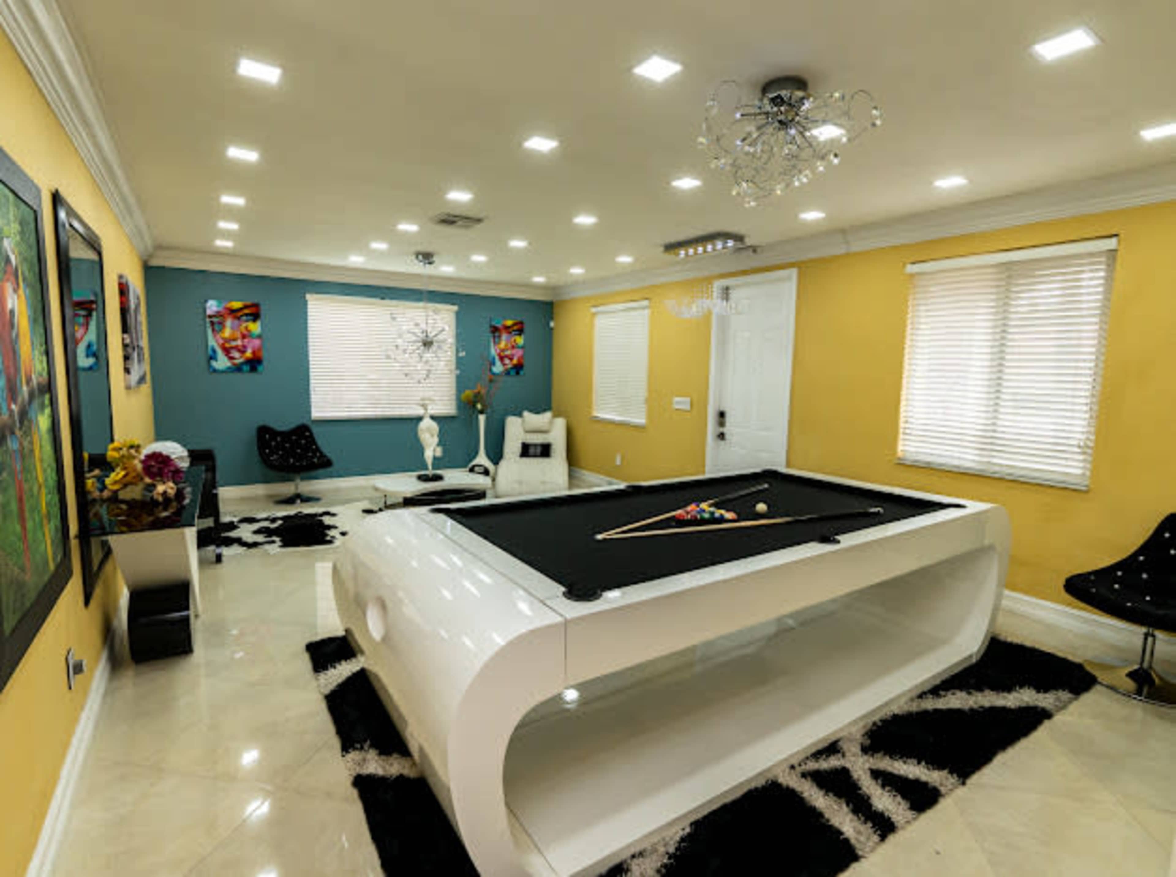 A modern game room with a sleek white pool table, surrounded by colorful artwork and contemporary furniture against a two-tone wall.