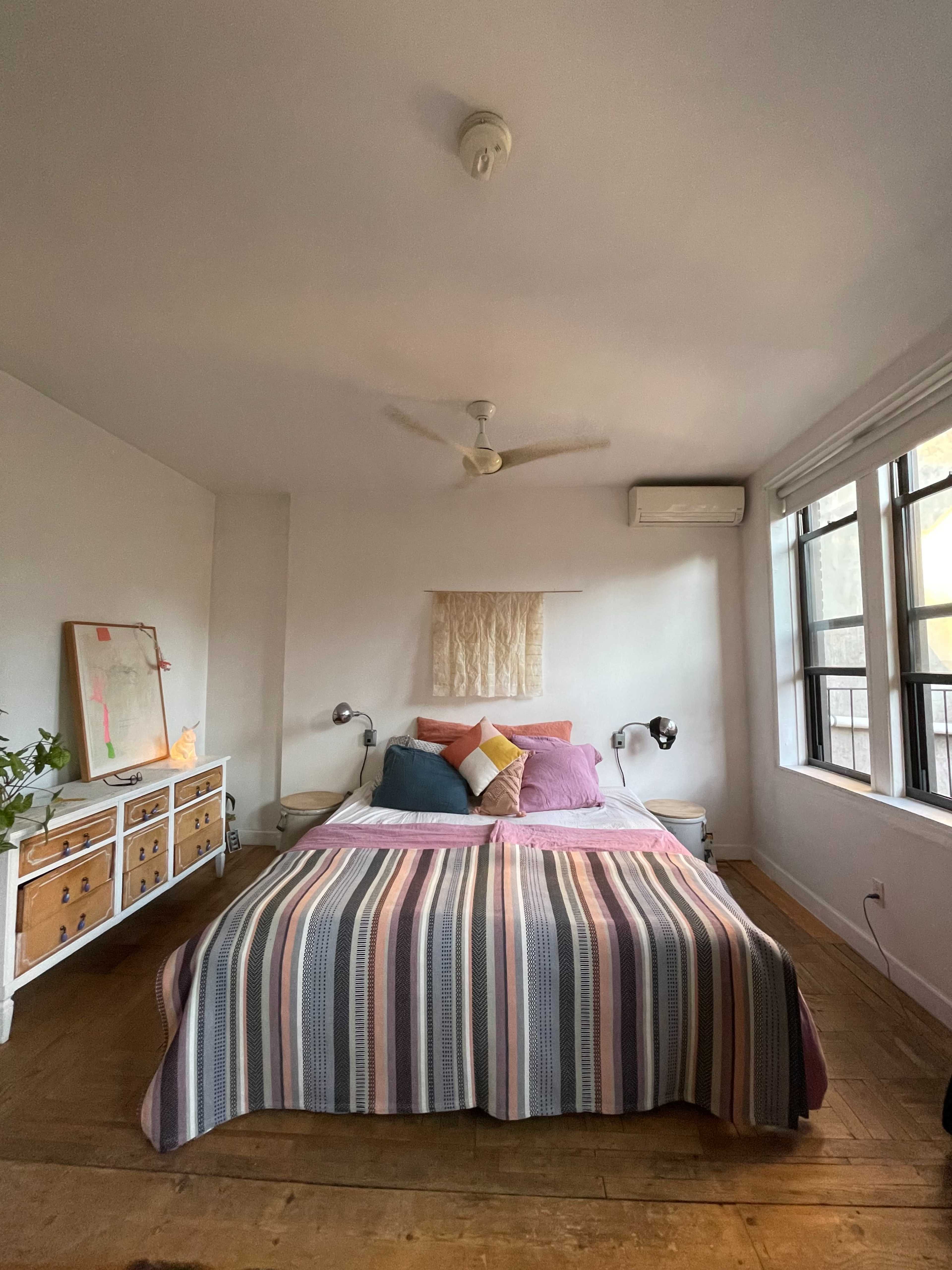 The image shows a neatly arranged bedroom with a bed dressed in striped linens, a wooden dresser, and large windows allowing natural light.