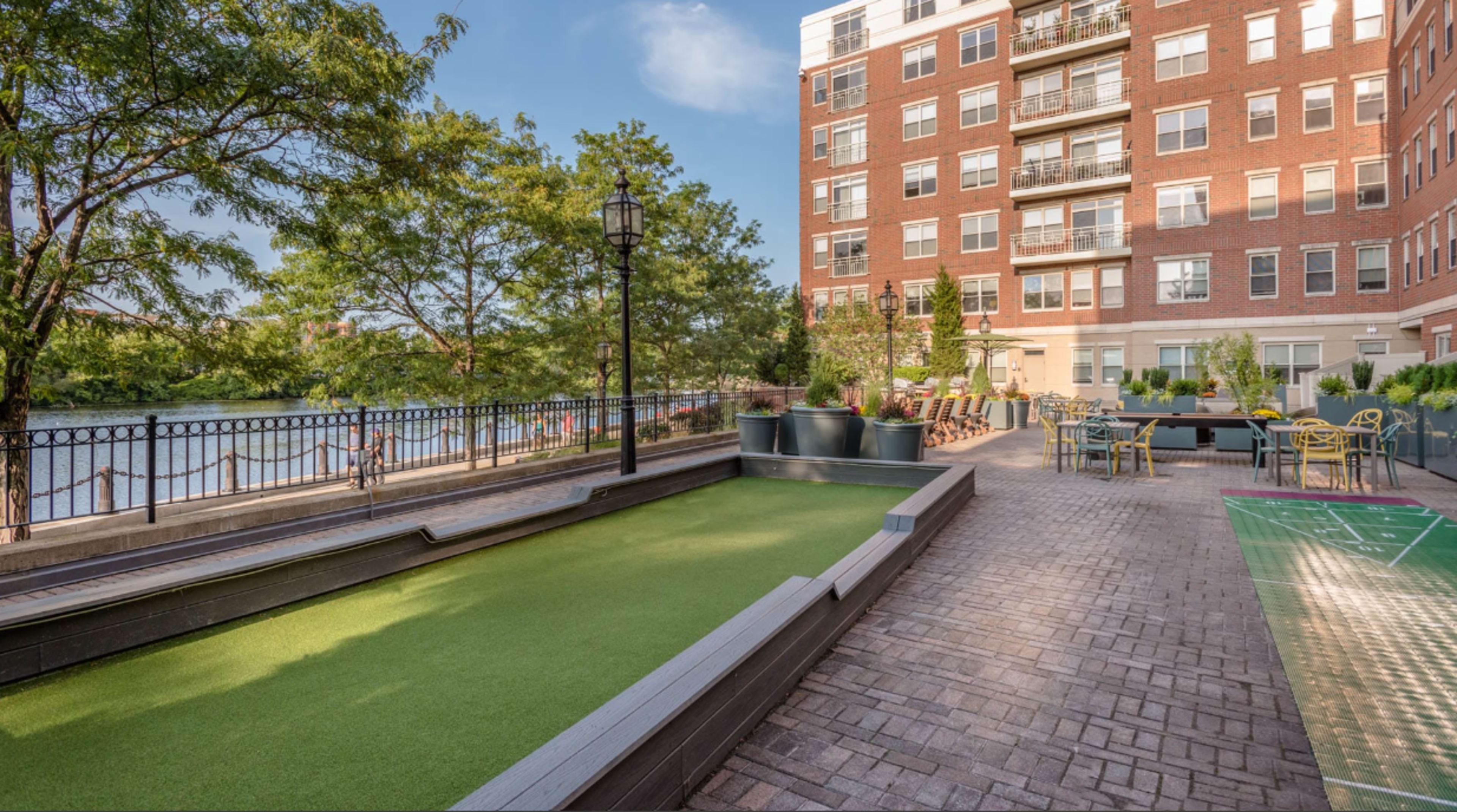 The image shows a landscaped outdoor area with a bocce ball court, seating, and a view of a river alongside a residential brick building.