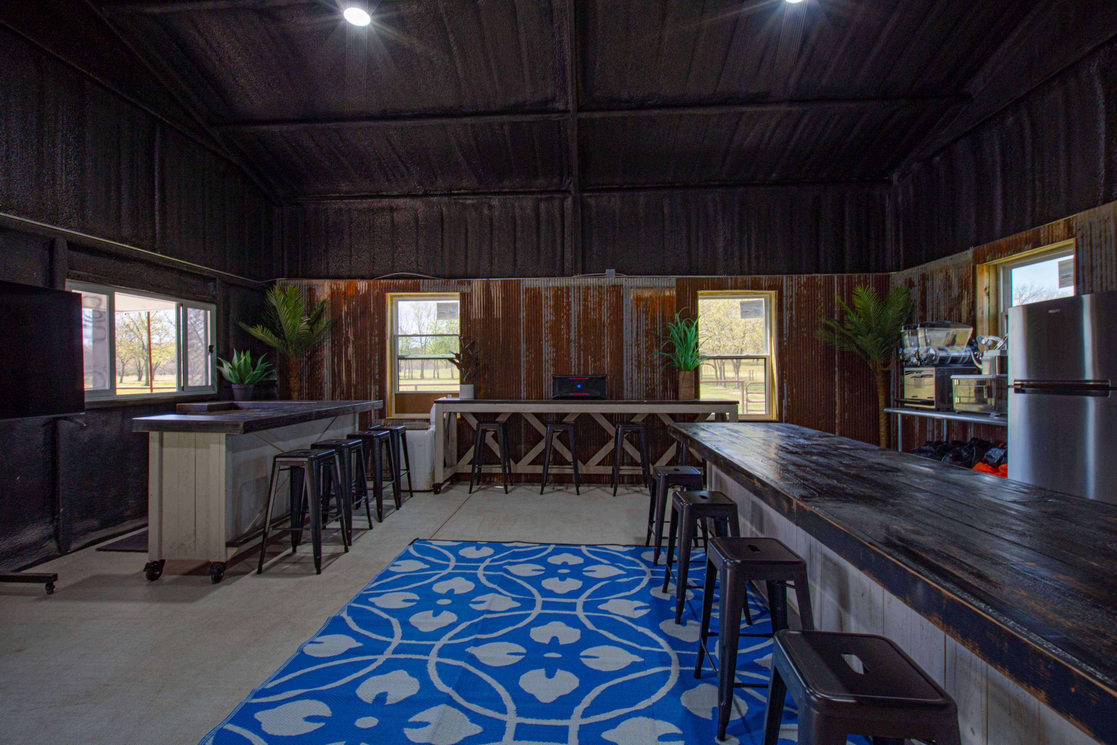 The image shows a spacious interior with a large patterned blue rug, rectangular tables, metal stools, and windows allowing natural light to enter.