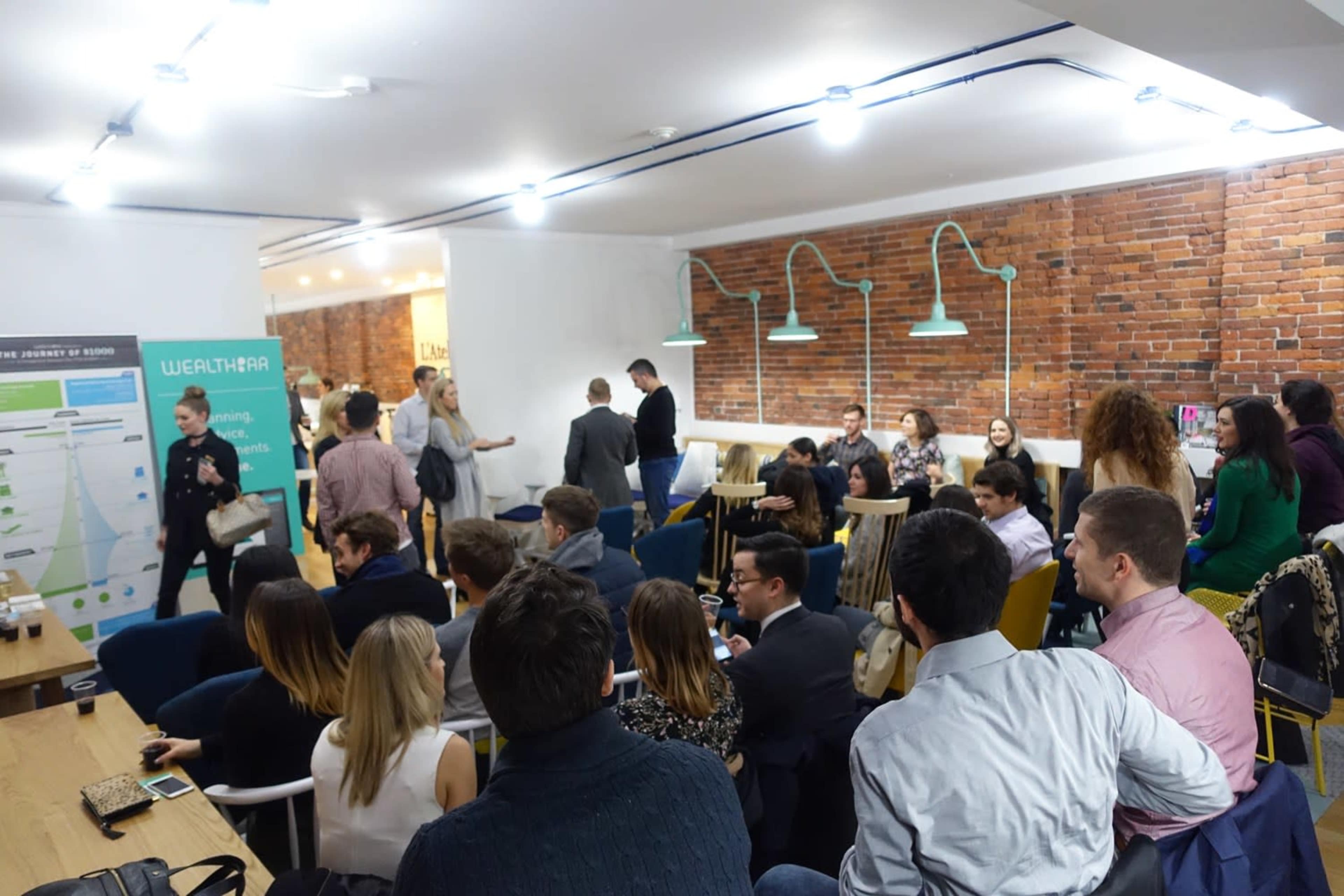 A group of people is seated and standing in a modern conference space with exposed brick walls and overhead lighting, engaged in conversation and watching a presentation.