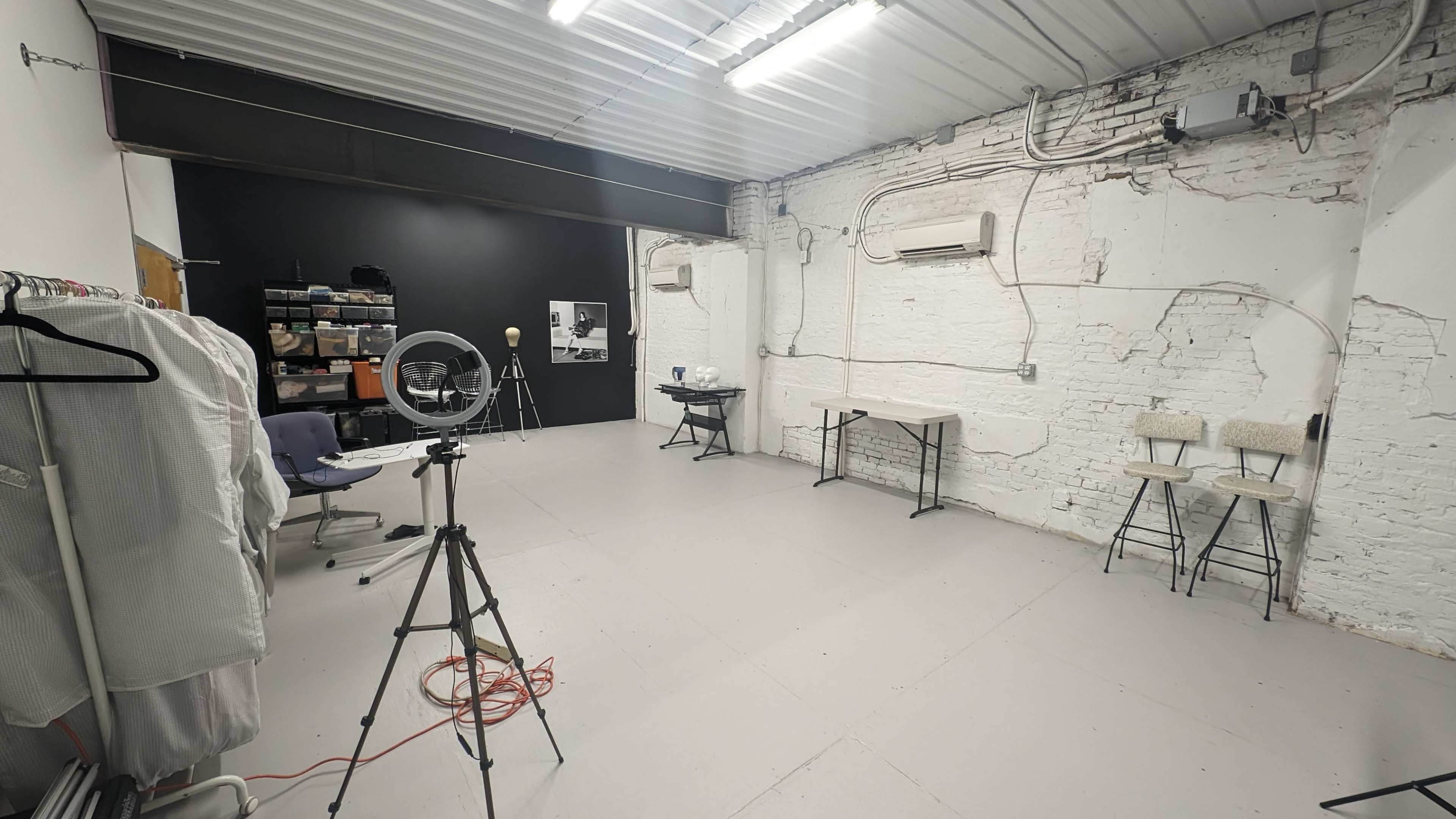 The image shows a spacious, empty studio with white walls, a black backdrop, various lighting equipment, and tables arranged for use.