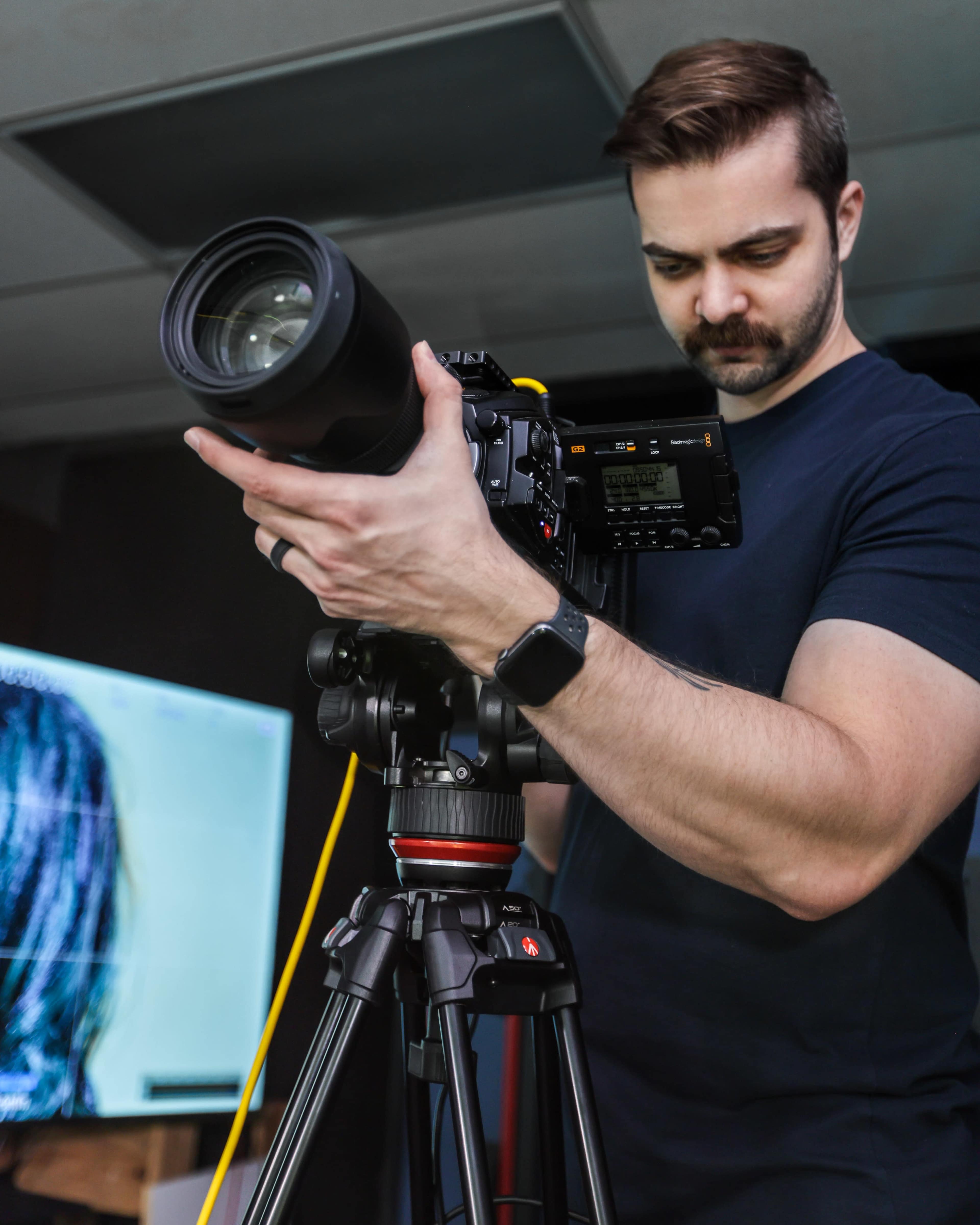 A photographer adjusts his camera on a tripod while focusing on a monitor displaying an image.
