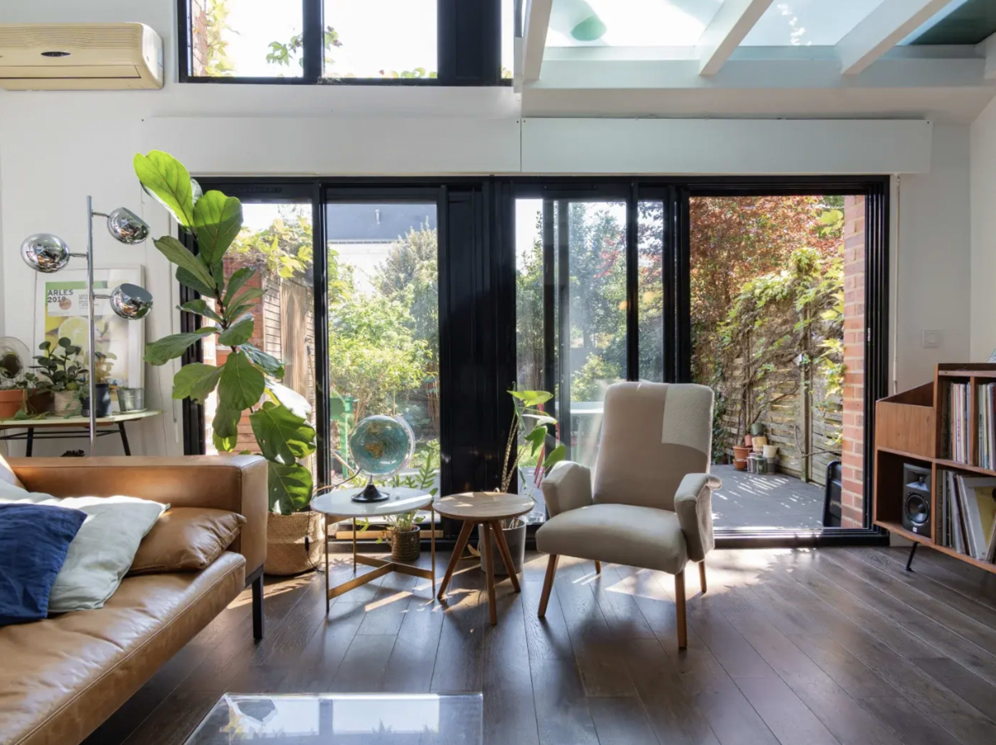 The image shows a modern living room with a brown leather sofa, a light armchair, large windows overlooking a garden, and various indoor plants.
