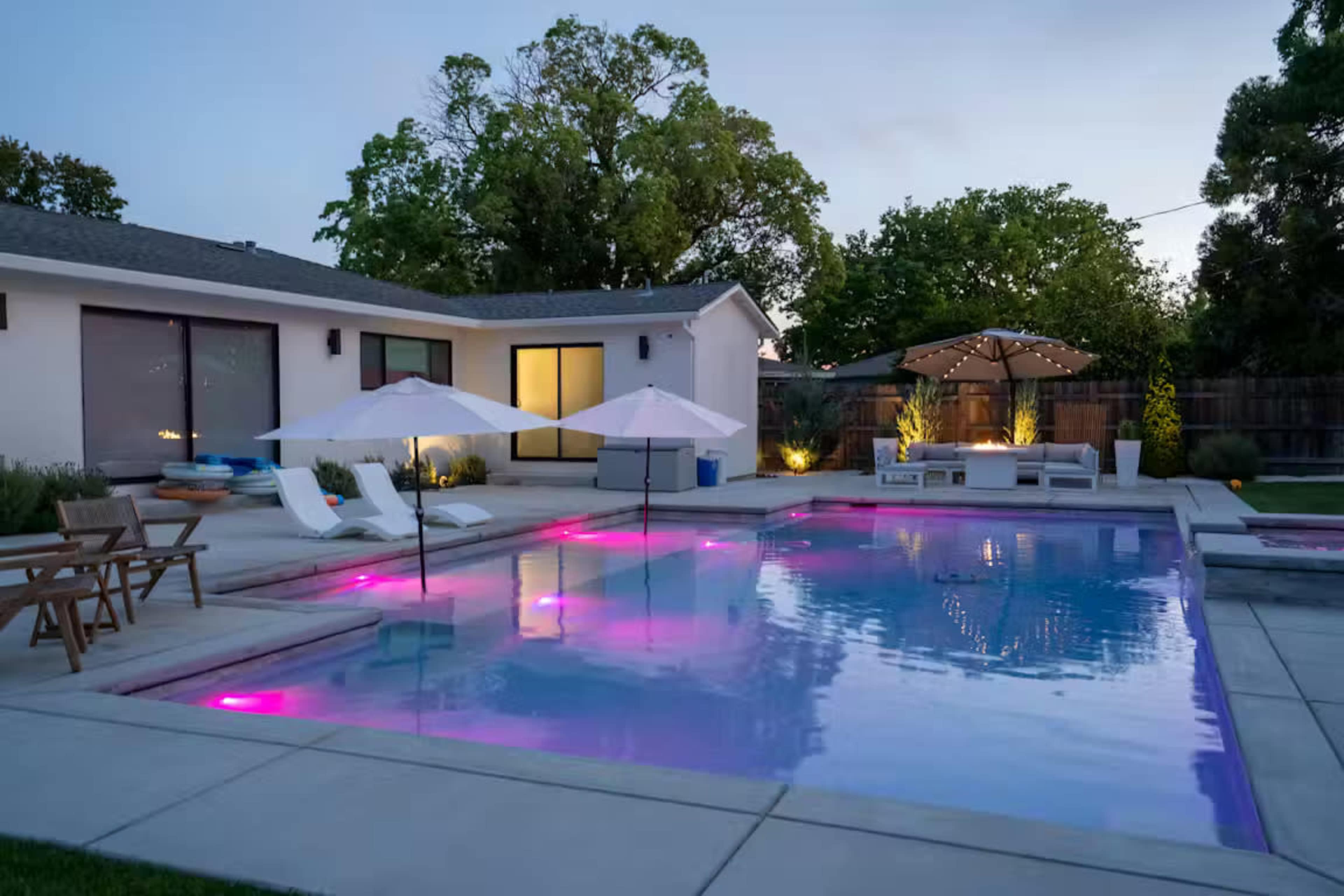 The image shows a modern backyard with a swimming pool featuring colorful lighting, lounge chairs, and a covered seating area.