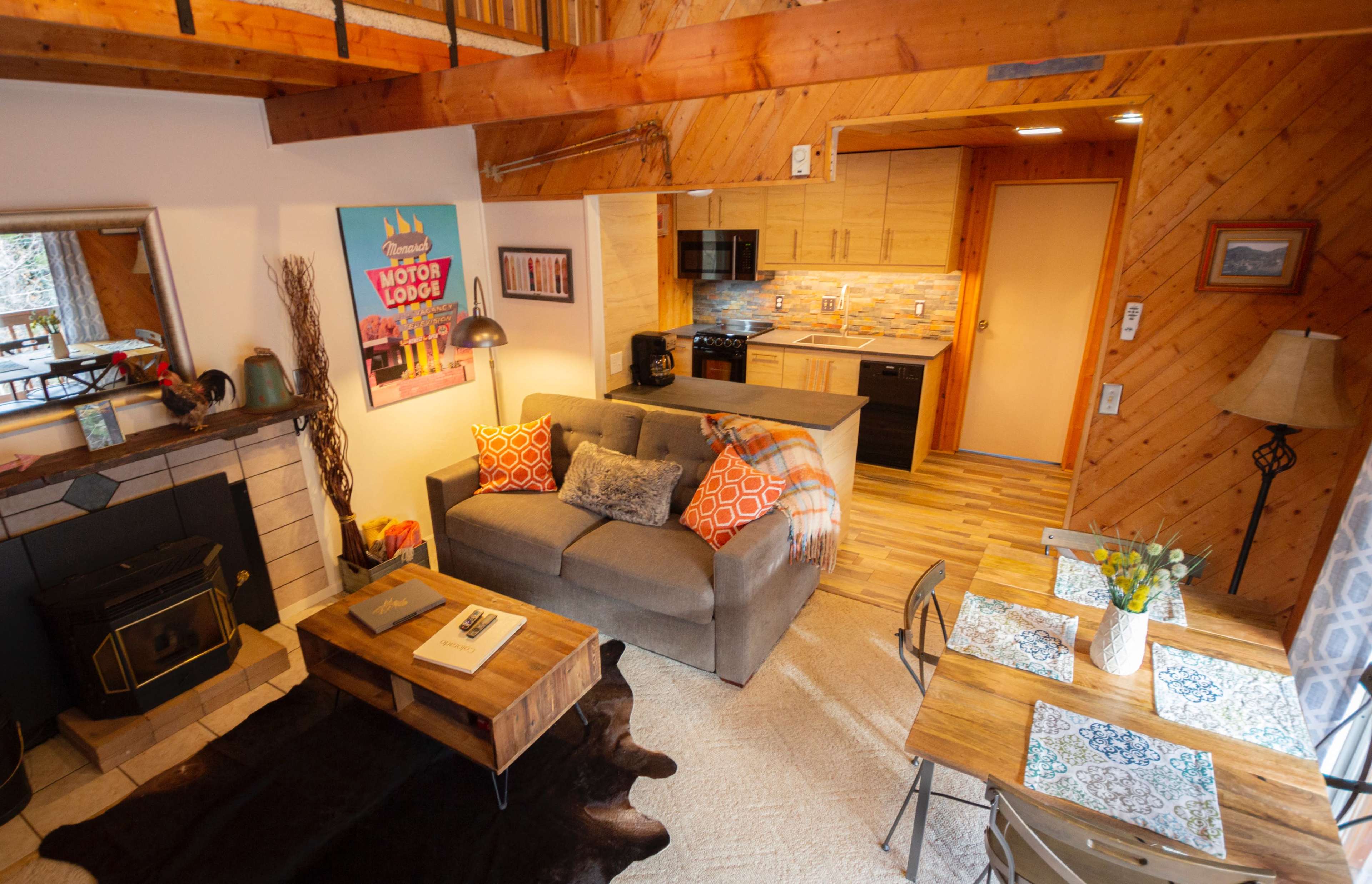 A cozy, wooden interior features a living area with a gray sofa, a coffee table, and a dining space with a floral centerpiece.