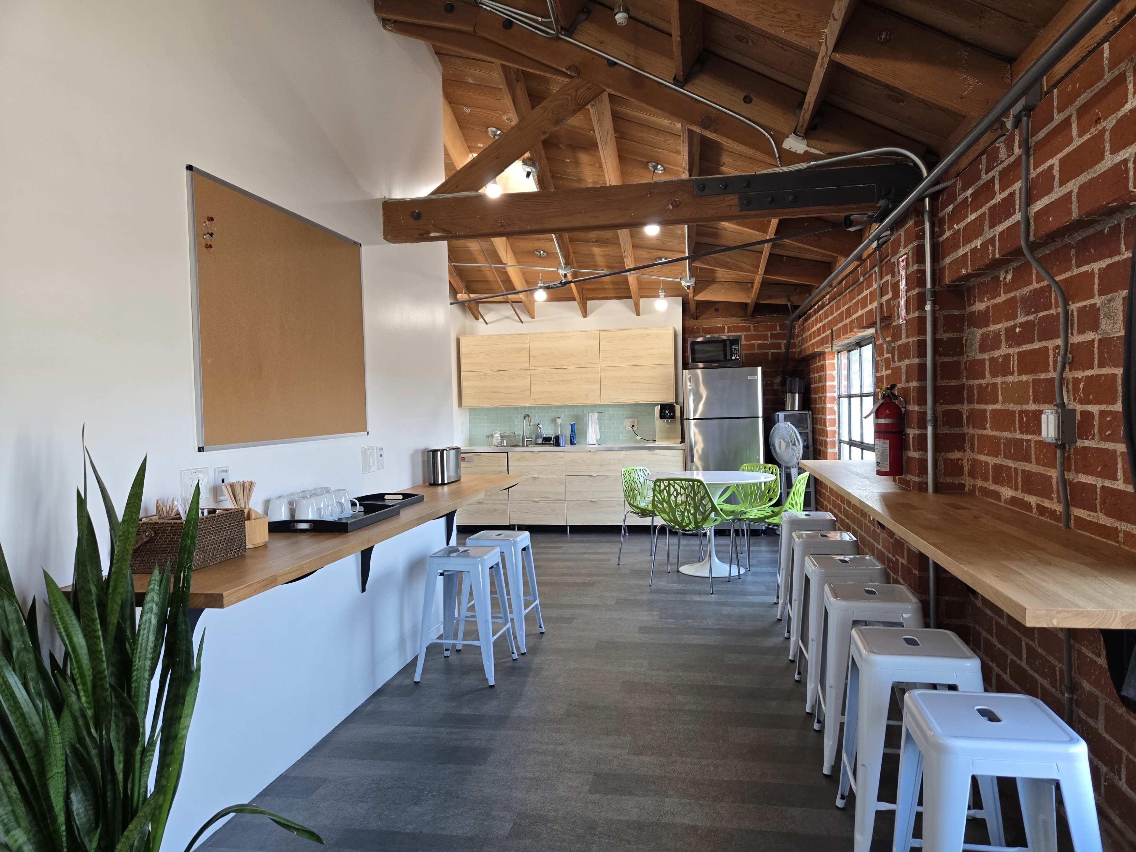 The image shows a modern kitchen area with exposed wooden beams, featuring a long countertop, metal stools, and a cooking space with light-colored cabinetry and appliances.