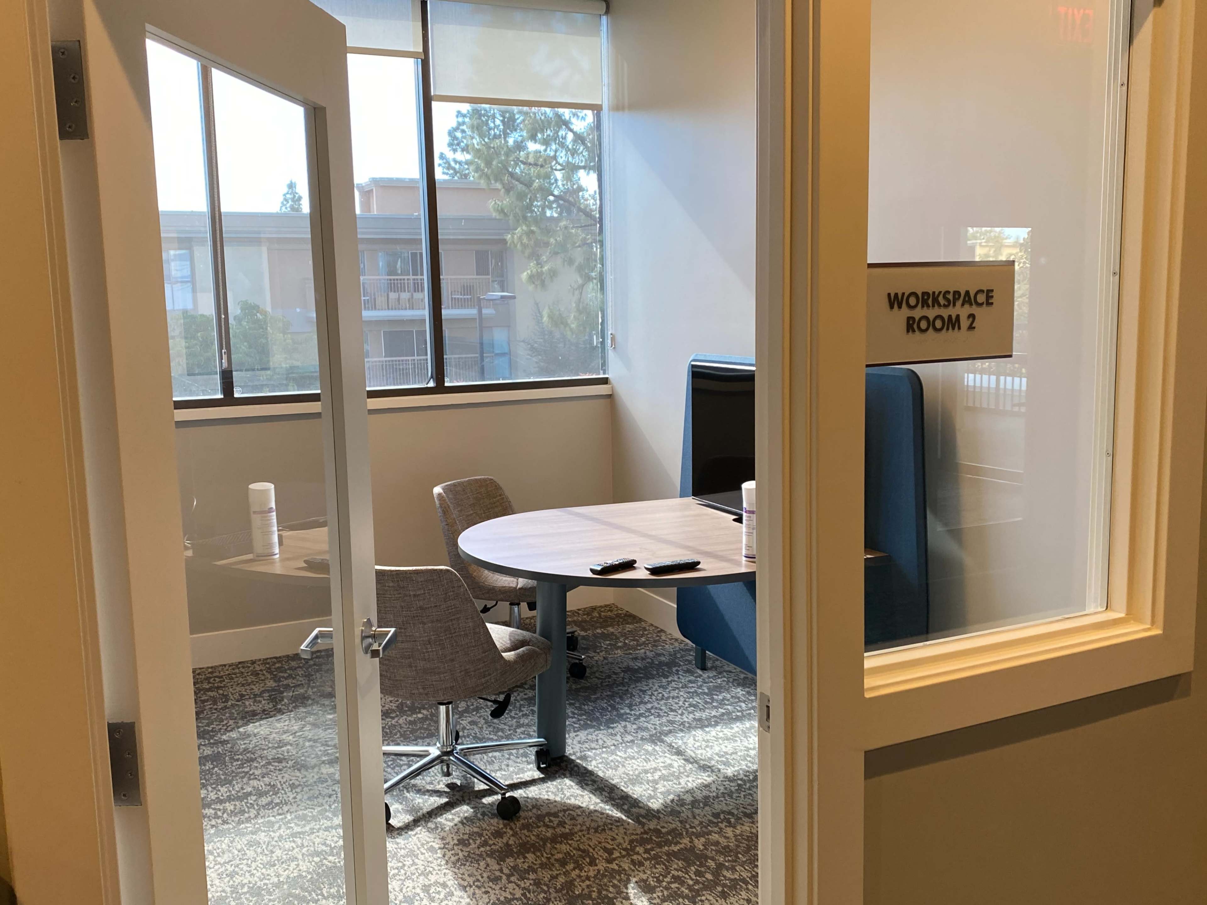 A small workspace with a round table and two chairs, visible through a glass door, in a well-lit room.