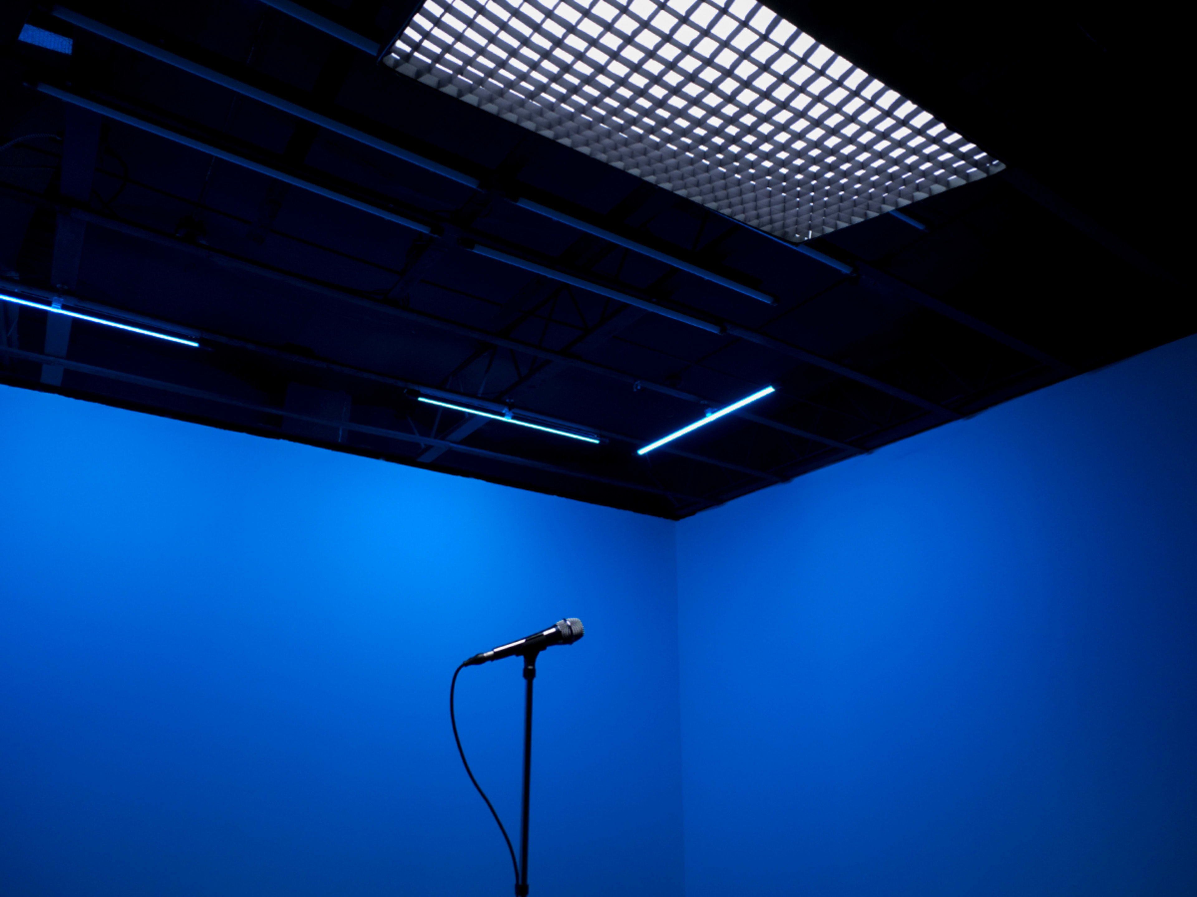 The image shows a minimalist stage setup with a single microphone on a stand against a blue background and a grid-patterned ceiling light.