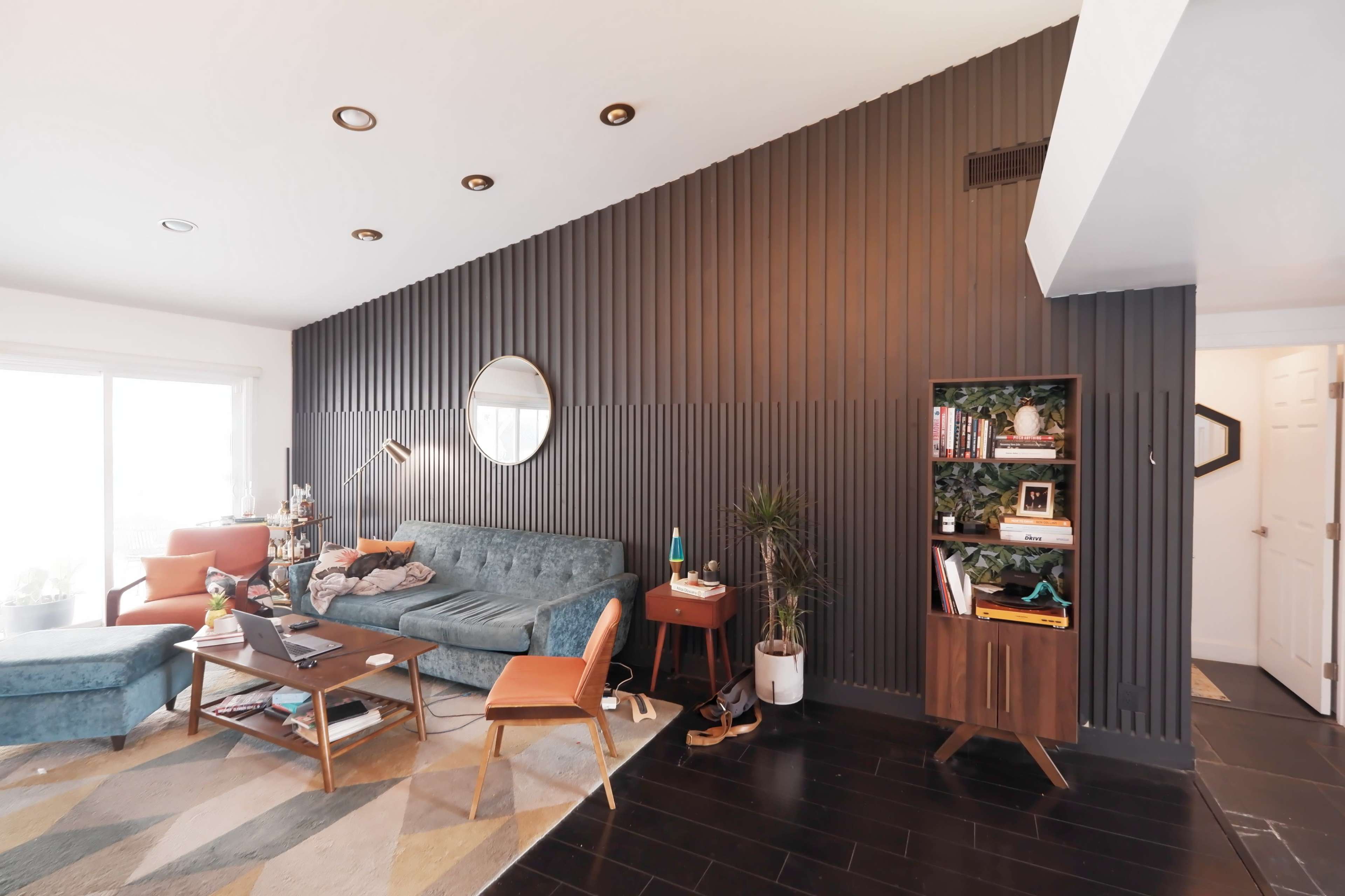 A modern living room featuring a teal sofa, a wooden coffee table, a bookshelf, and a dark paneled wall with a round mirror.