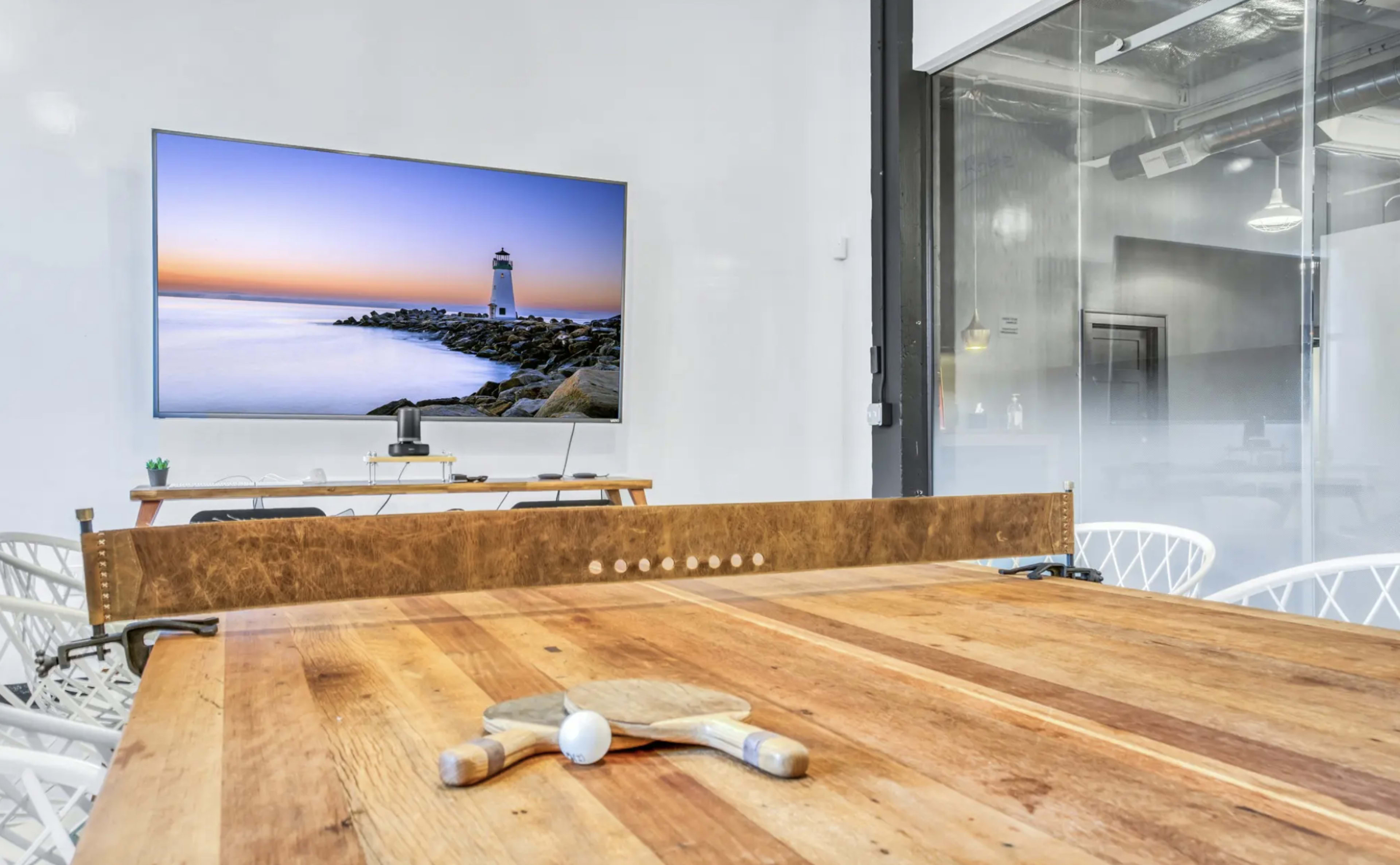 A wooden conference table with table tennis paddles and a ball is positioned in front of a large television displaying a coastal lighthouse scene.