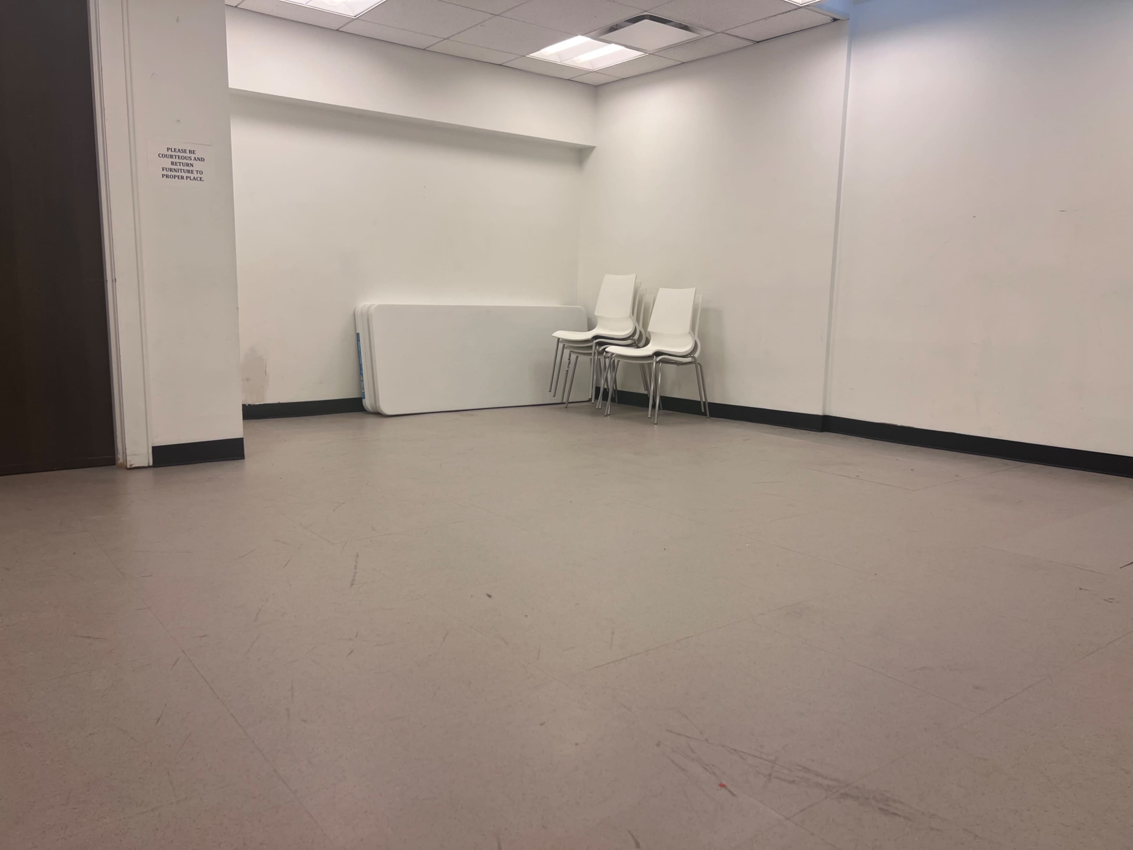 The image shows a large, empty room with a few white chairs stacked against a wall and a flat surface partially visible in the background.
