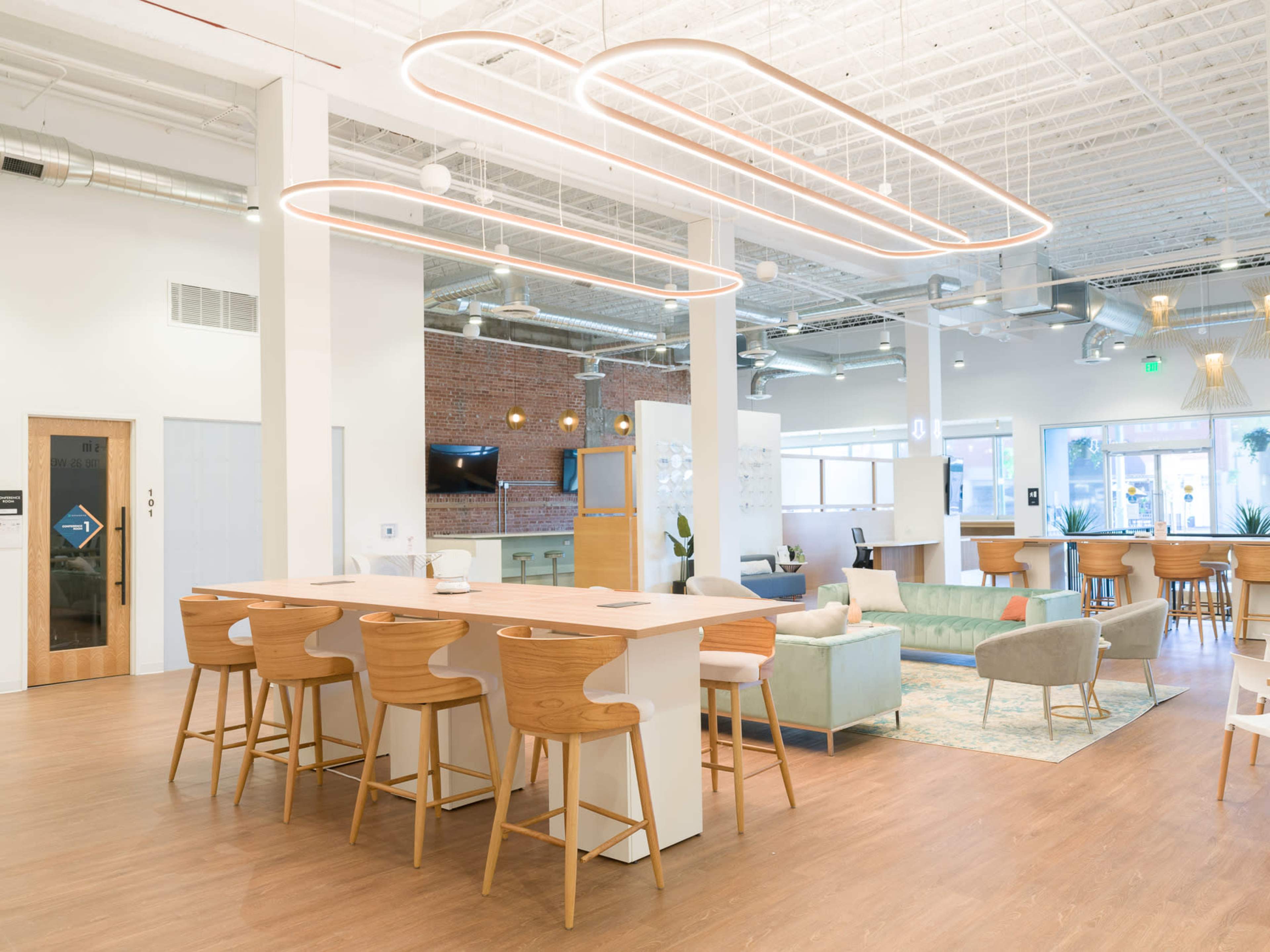The image shows a bright, modern co-working space with a central bar-style table, wooden chairs, and various seating areas arranged around the room.
