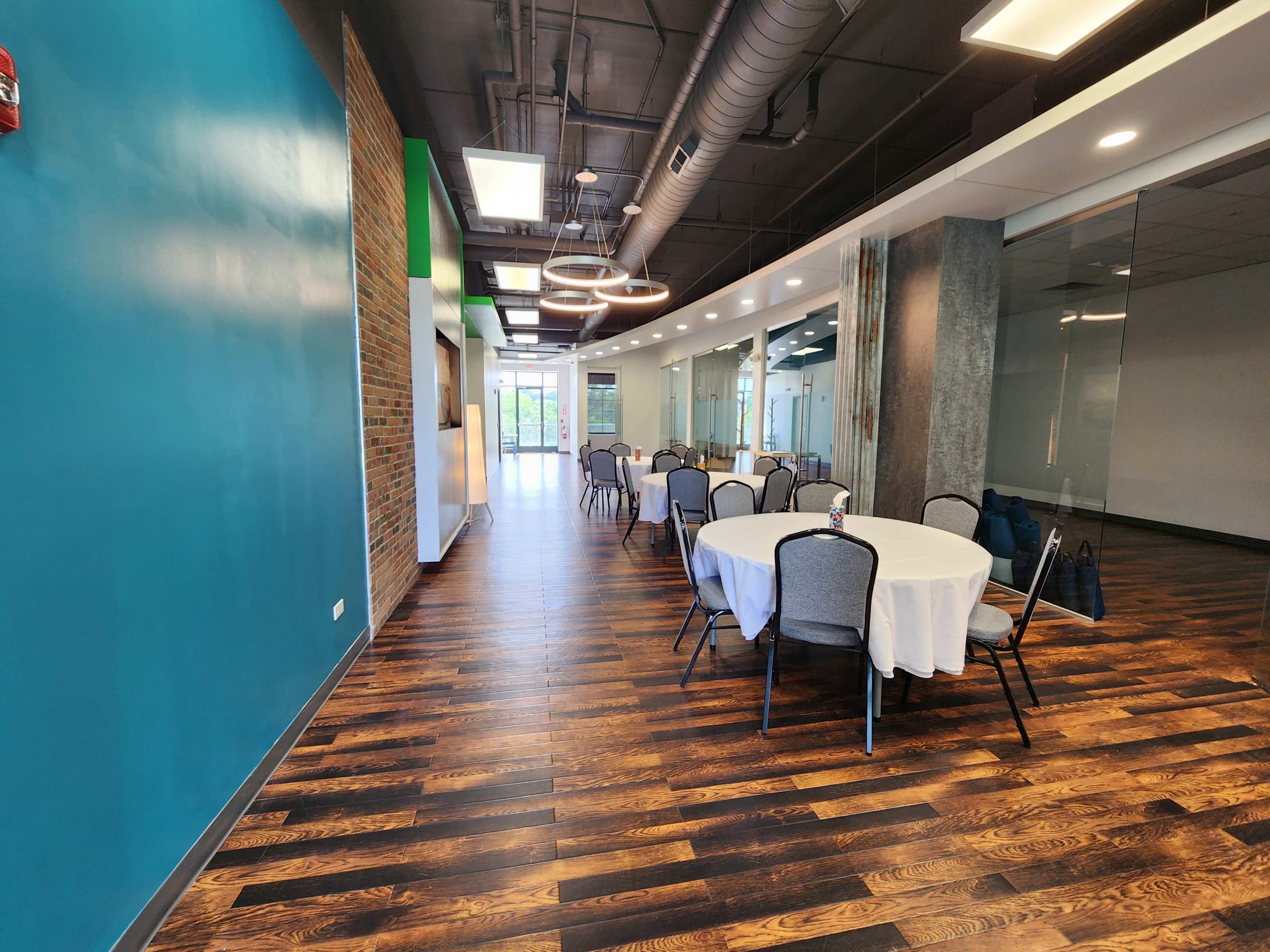 A well-lit interior space with round tables covered in white tablecloths, wooden flooring, and a teal accent wall.