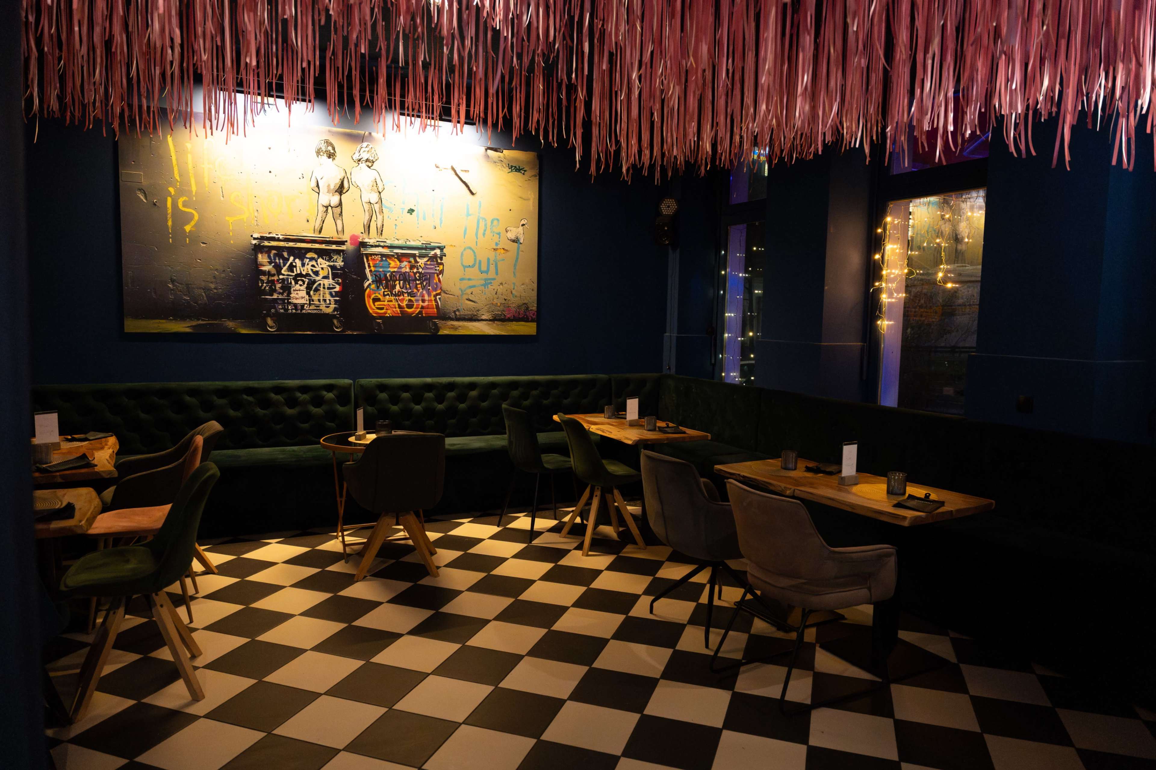 The image shows a modern restaurant interior featuring checkered flooring, upholstered seating, and a colorful artwork on the wall, with pink streamers hanging from the ceiling.