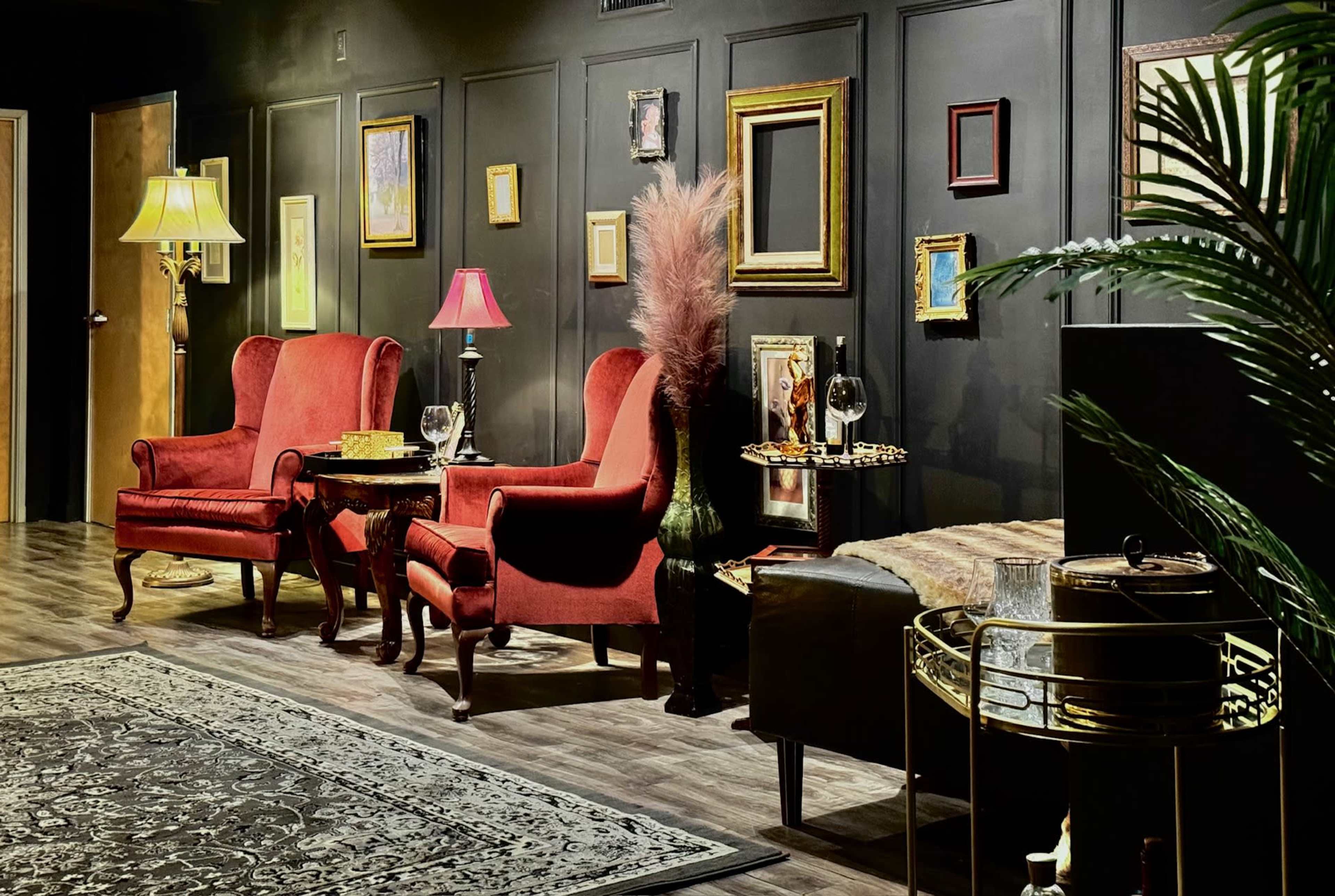 The scene features two red armchairs placed opposite each other, surrounded by various framed artworks on a dark wall, with lamps, a potted plant, and a decorative table nearby.