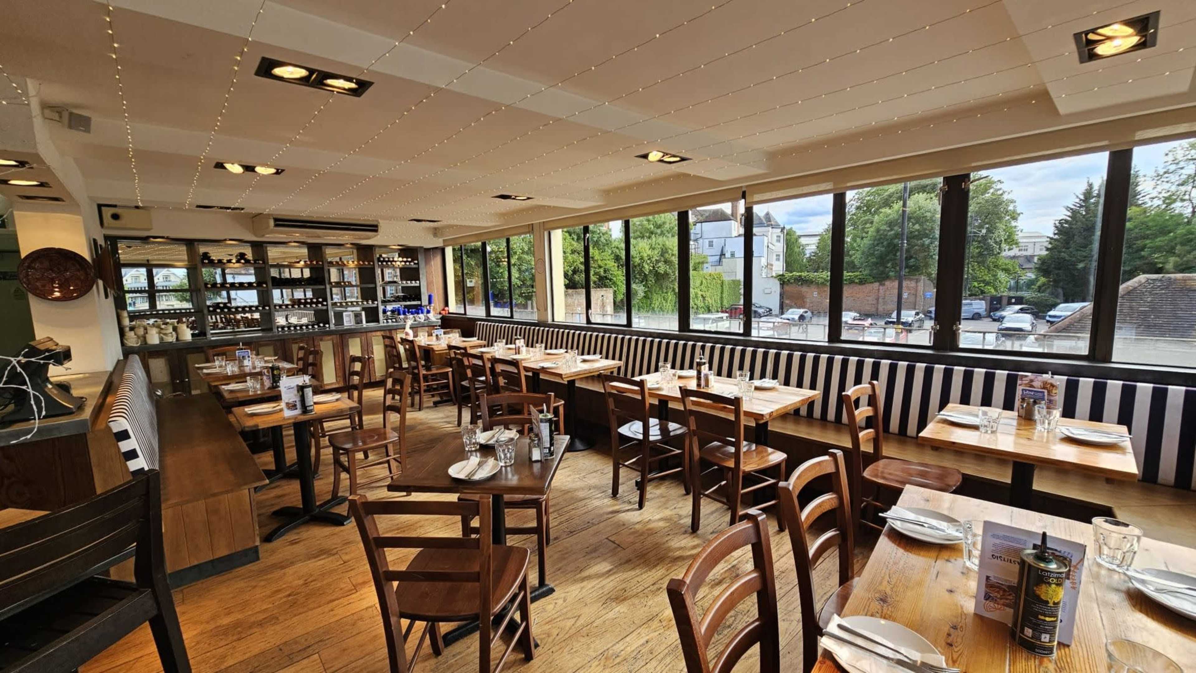 The image shows the interior of a restaurant with wooden tables and chairs, large windows allowing natural light, and a bar area in the background.