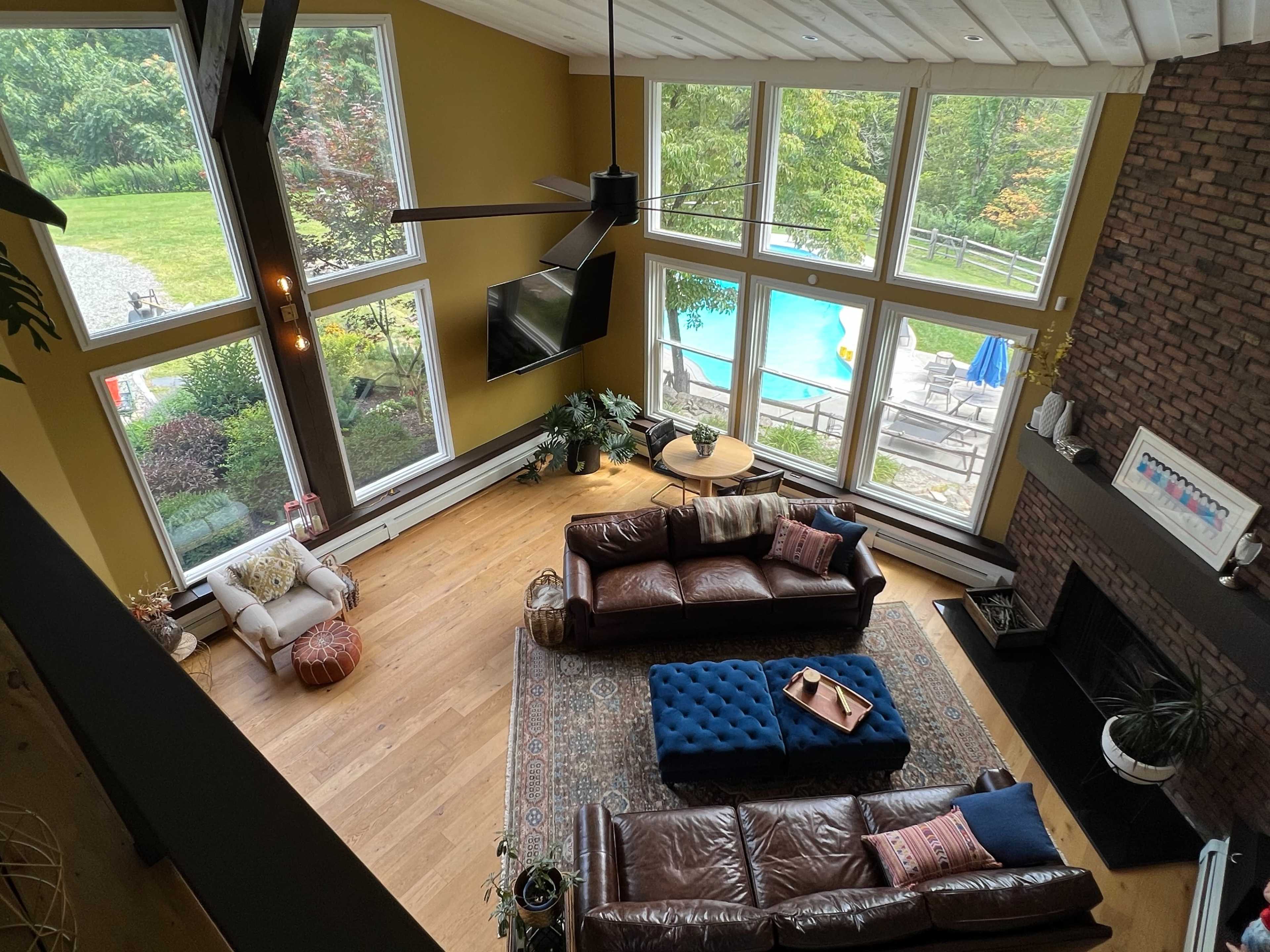 The image shows a spacious living room featuring large windows that overlook a pool area, with a mix of leather sofas, a coffee table, and a patterned area rug.