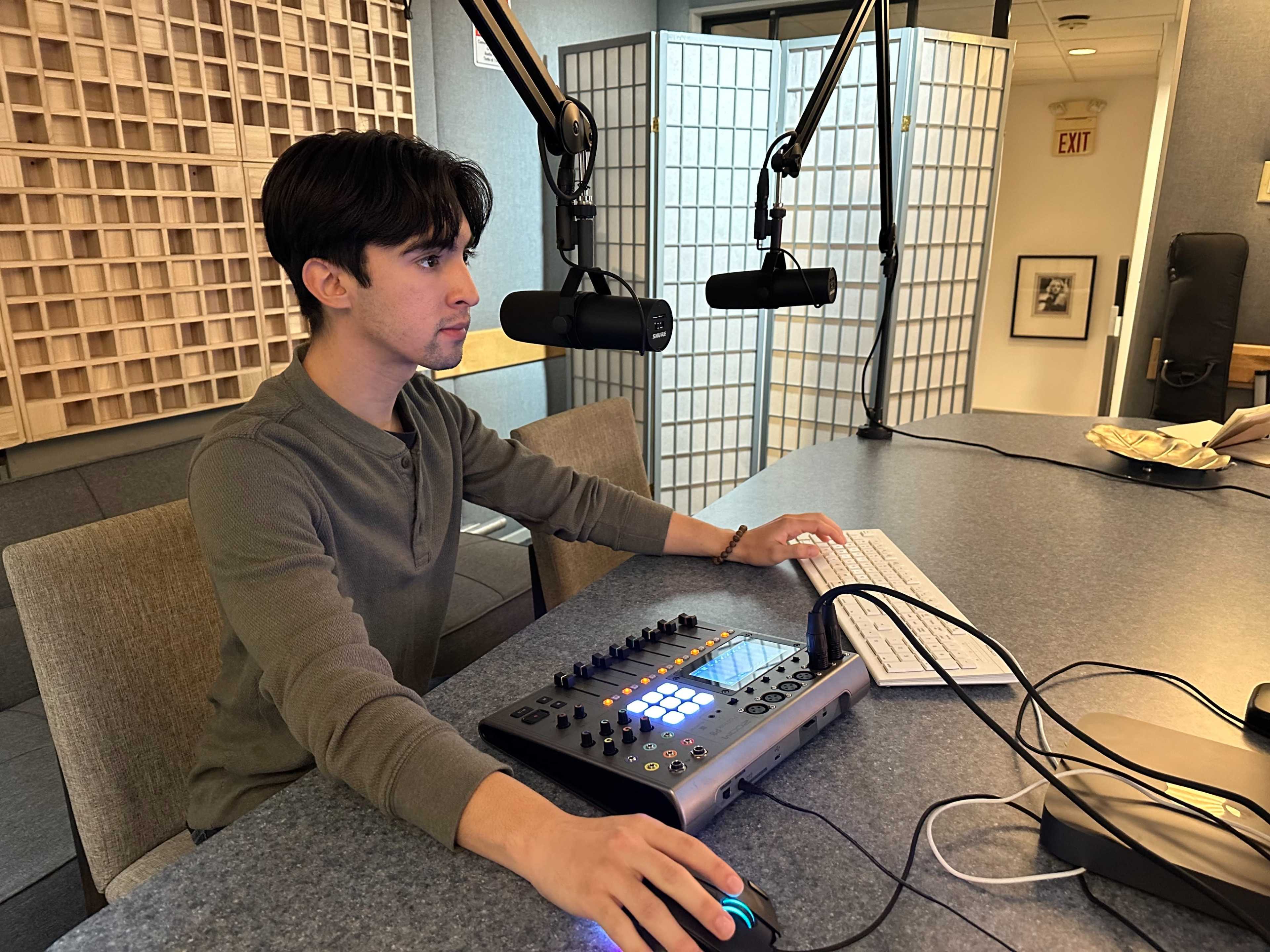 A young man sits at a desk in a recording studio, operating audio equipment while facing two microphones.