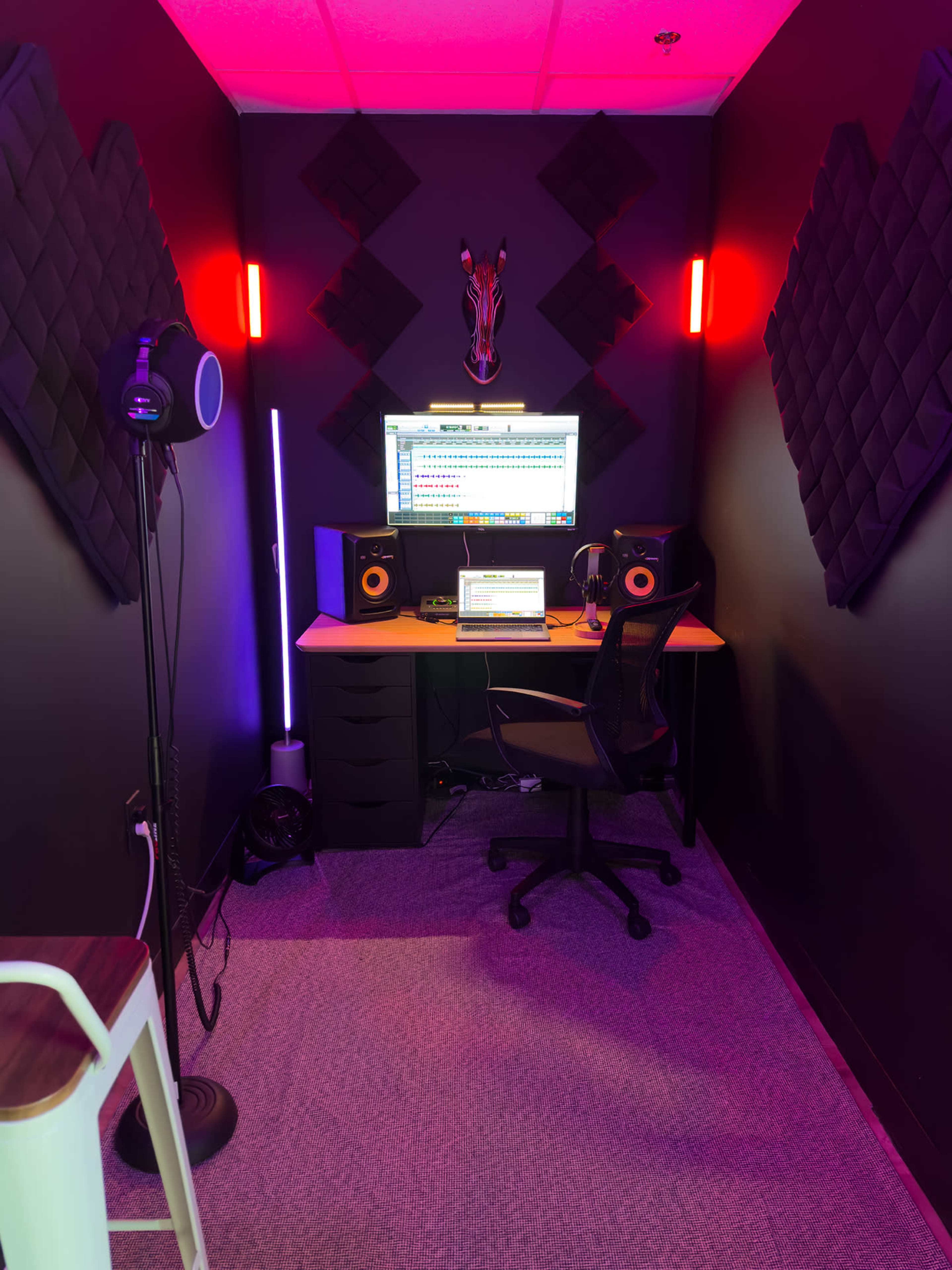 A small recording studio with a desk, computer monitor, speakers, and soundproofing panels on the walls, illuminated by colorful lighting.