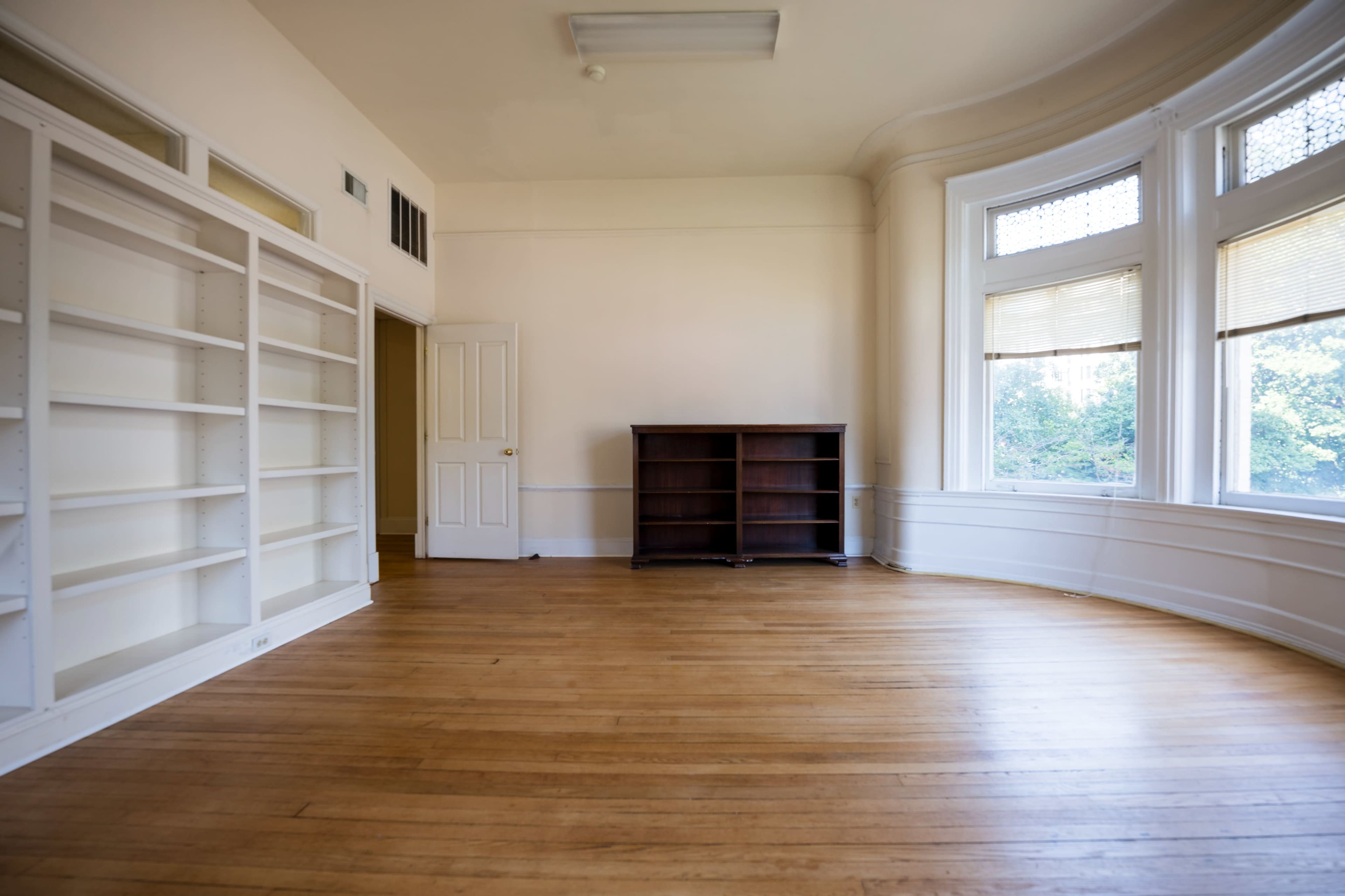 The image shows a vacant room with wooden floors, empty built-in shelves, and a dark wooden cabinet against a curved wall with windows.