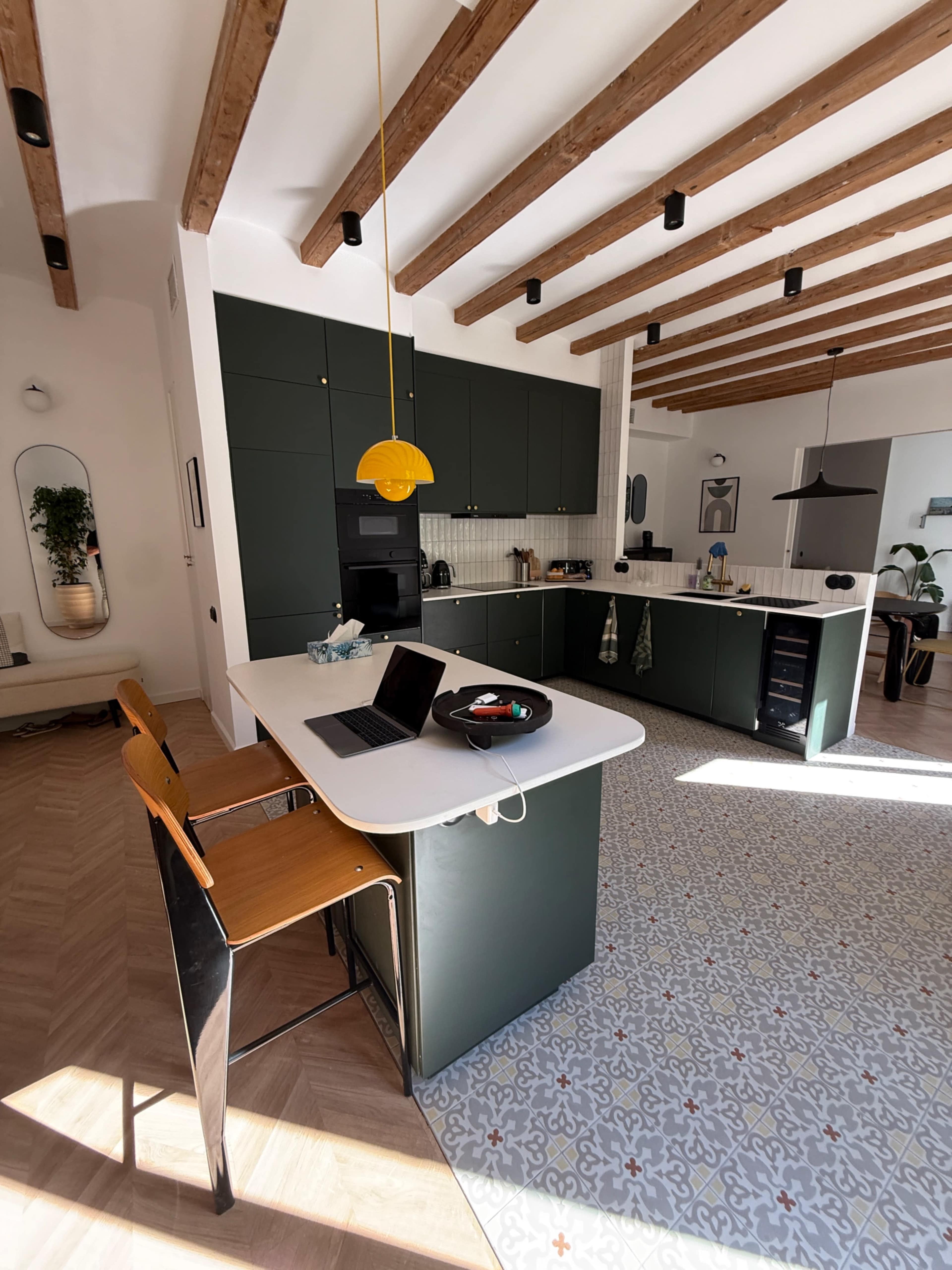 The image shows a modern kitchen with a central island, wooden ceiling beams, green cabinets, and a patterned floor.