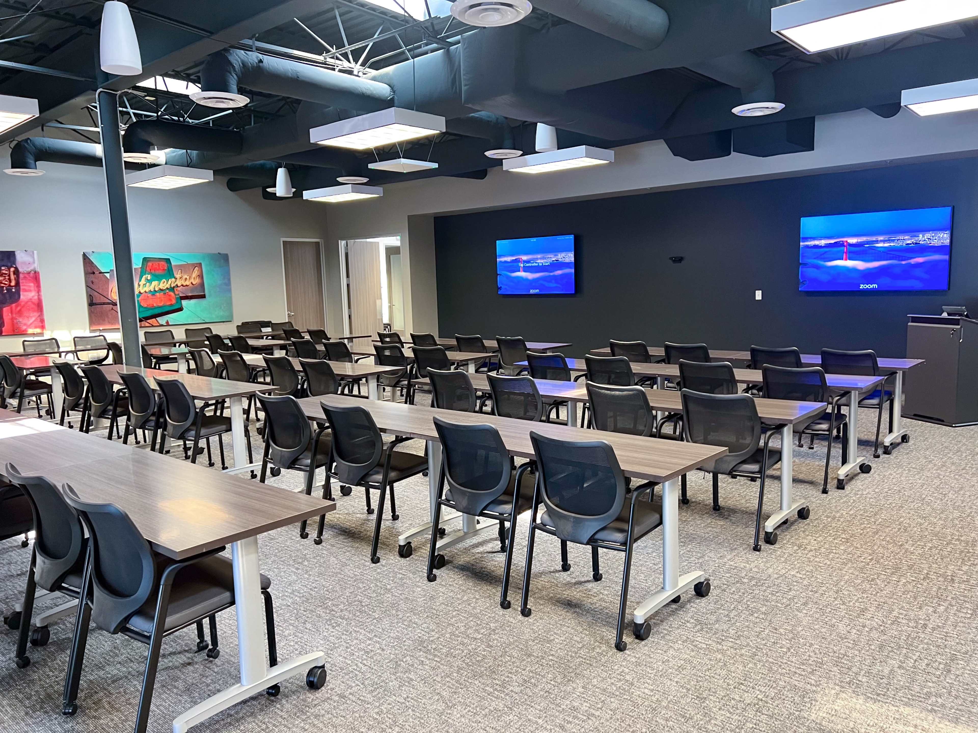 The image shows a modern classroom setup with multiple rows of tables and chairs, two large screens on the walls, and a neutral-colored carpet.