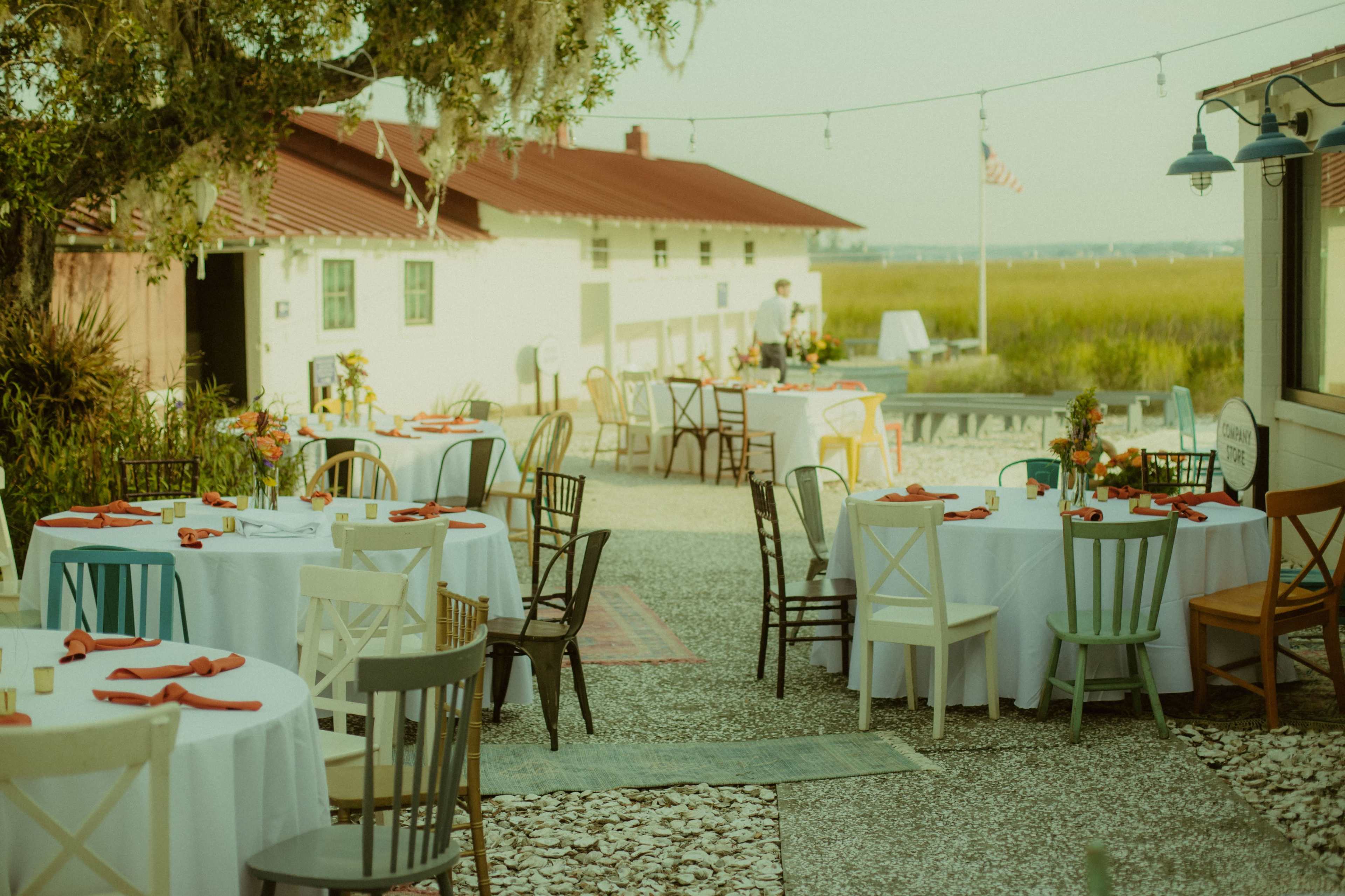 An outdoor dining area is set with various tables and chairs, decorated with flowers and tableware, against a backdrop of water and a clear sky.