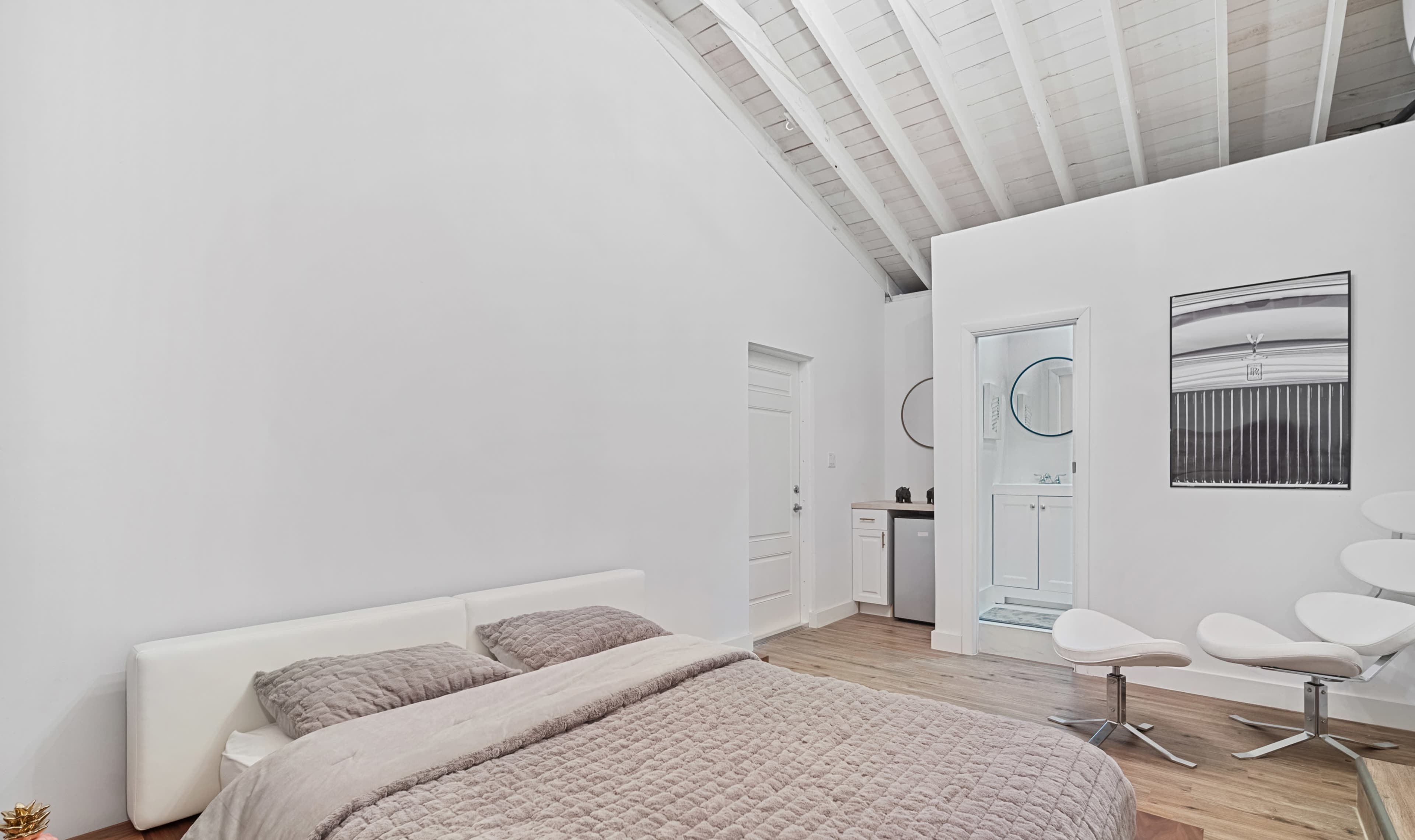 The image shows a bedroom with a bed covered in a light quilt, wooden flooring, and a doorway leading to a small bathroom.