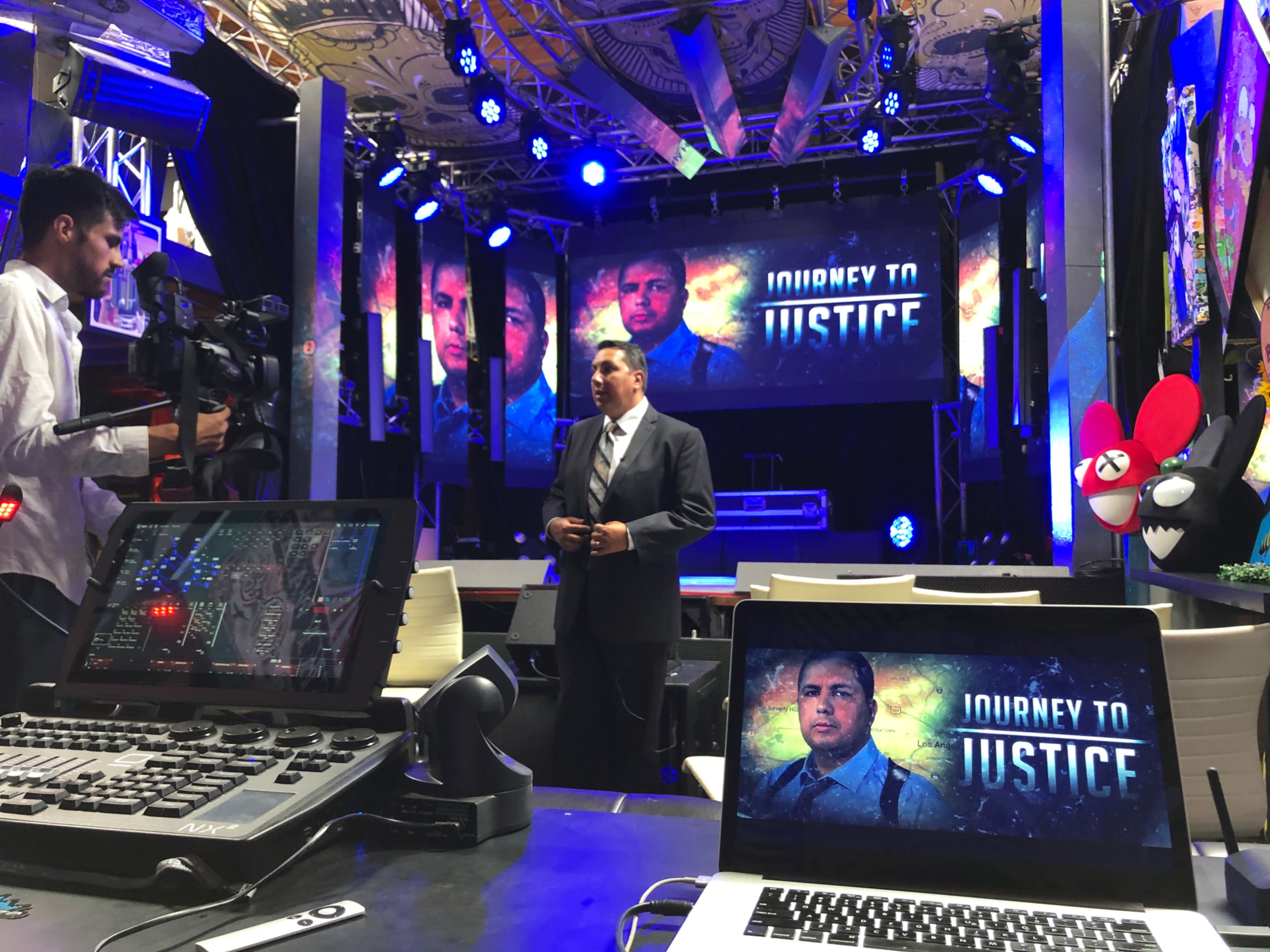A man speaks on stage in front of multiple screens displaying "Journey to Justice" while a camera operator films the presentation.