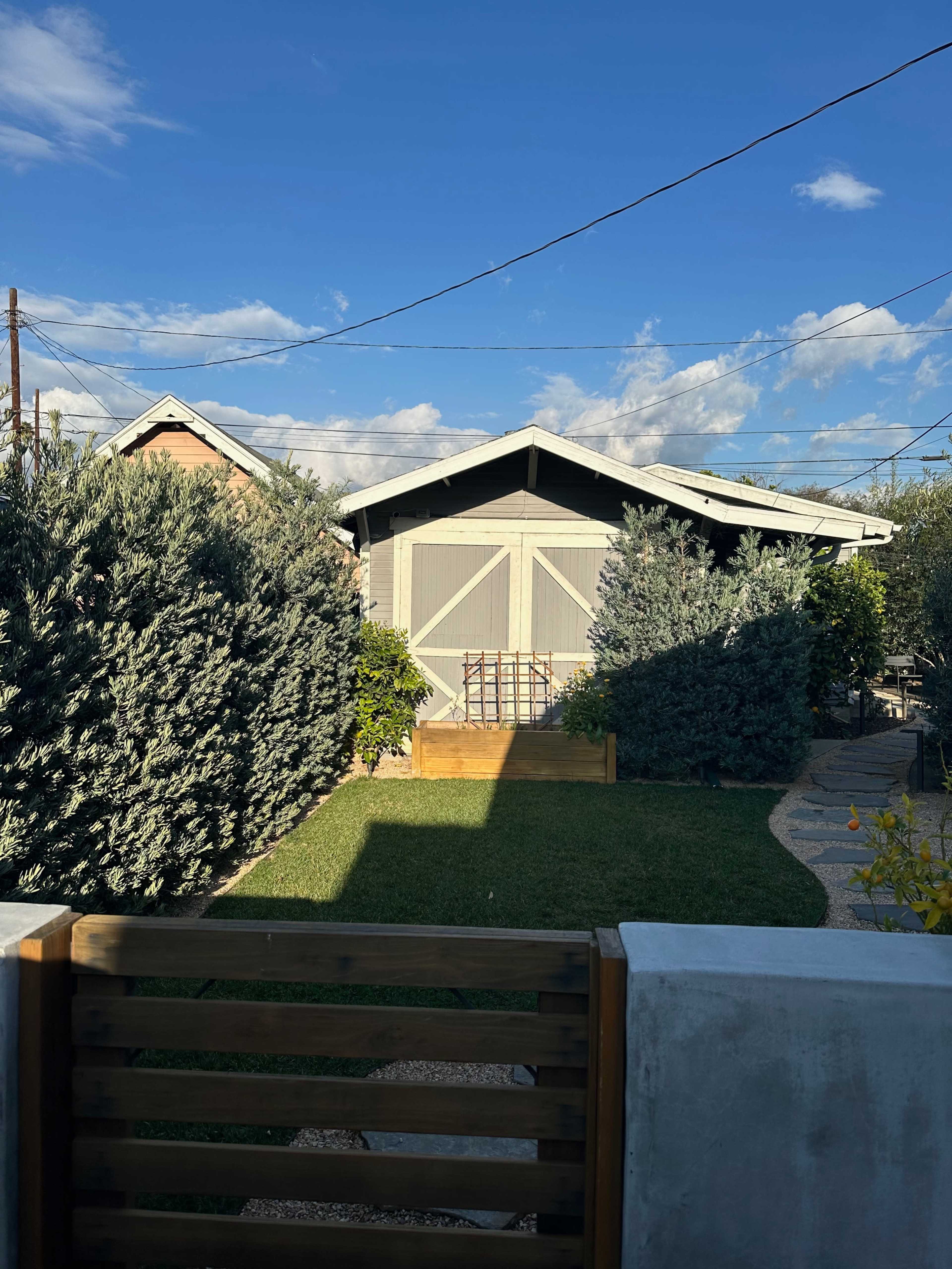 A wooden shed with a sloped roof is surrounded by tall shrubs and grass under a blue sky with scattered clouds.