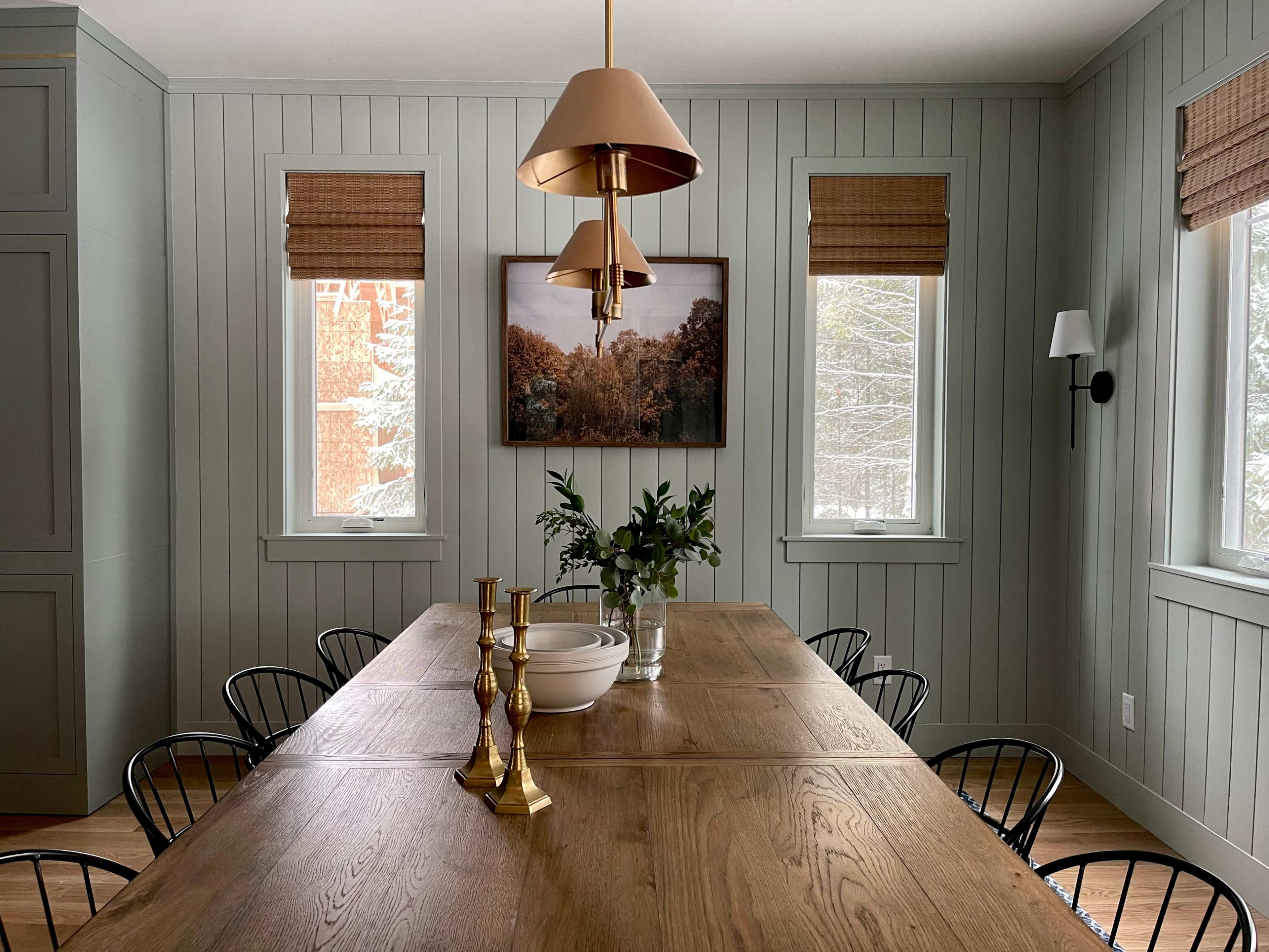 A spacious dining room features a long wooden table surrounded by black chairs, with a large pendant light overhead and wall art displayed between two windows.