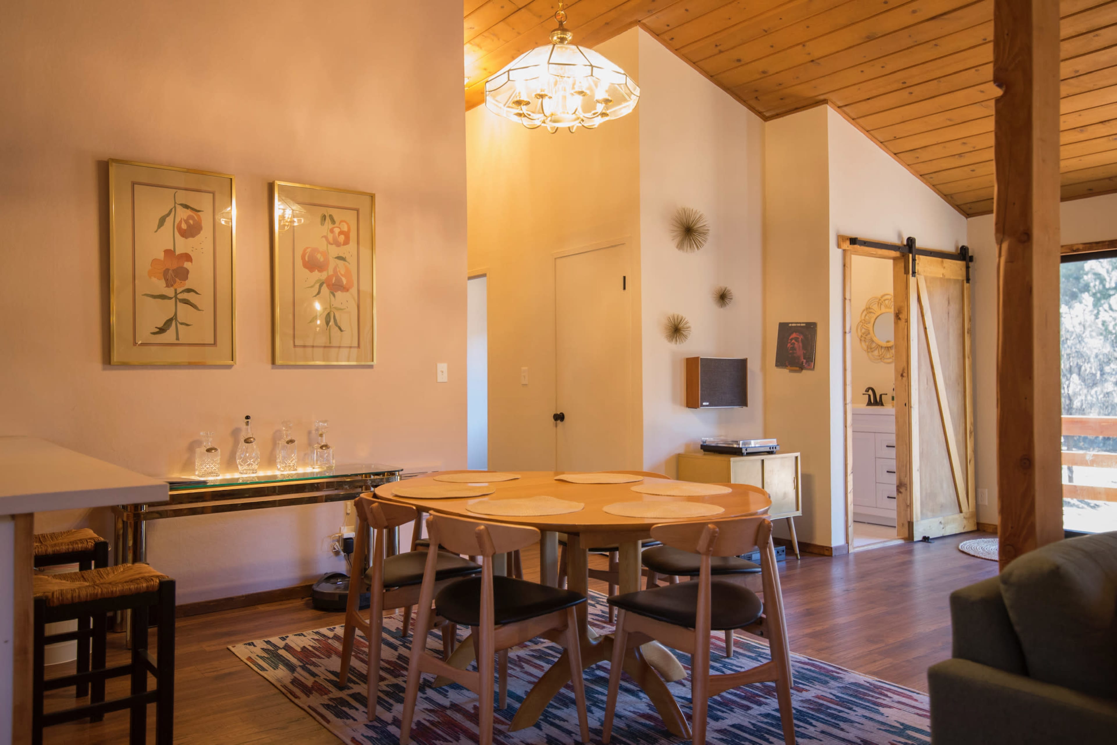 A well-lit dining area features a round table with wooden chairs, framed floral artwork on the walls, and a sliding door leading to a balcony.