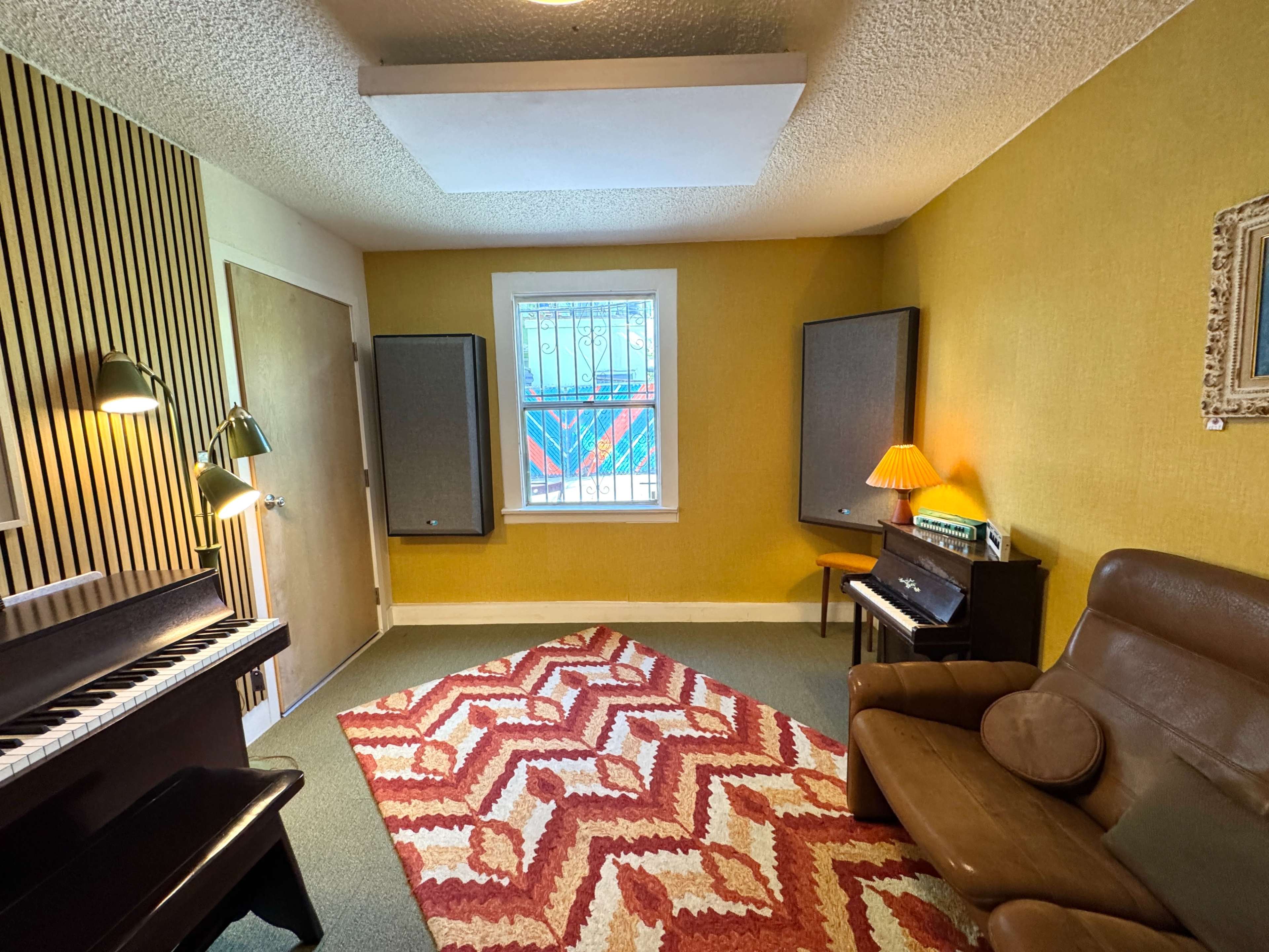 A small room features a piano, a brown leather chair, patterned rug, and two large speakers, with a window overlooking an exterior view.