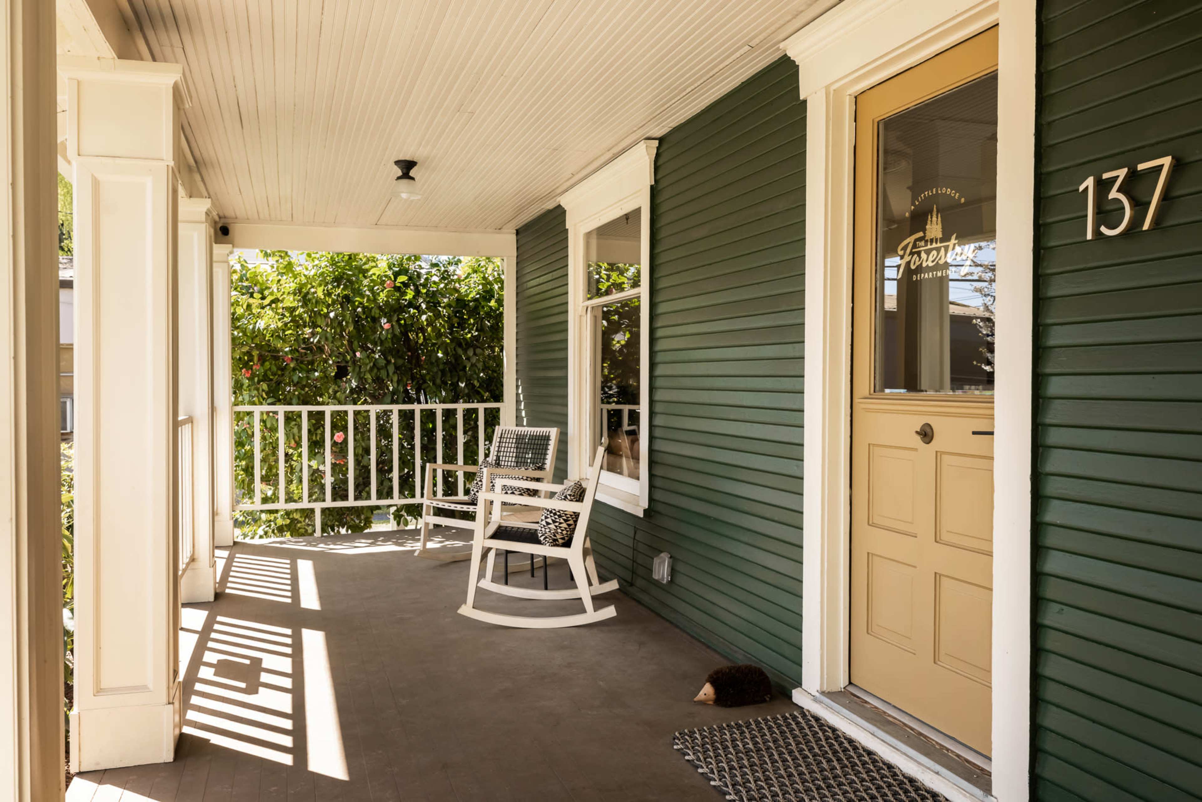 The image shows a green porch with a rocking chair, a door numbered 137, and a view of greenery outside.