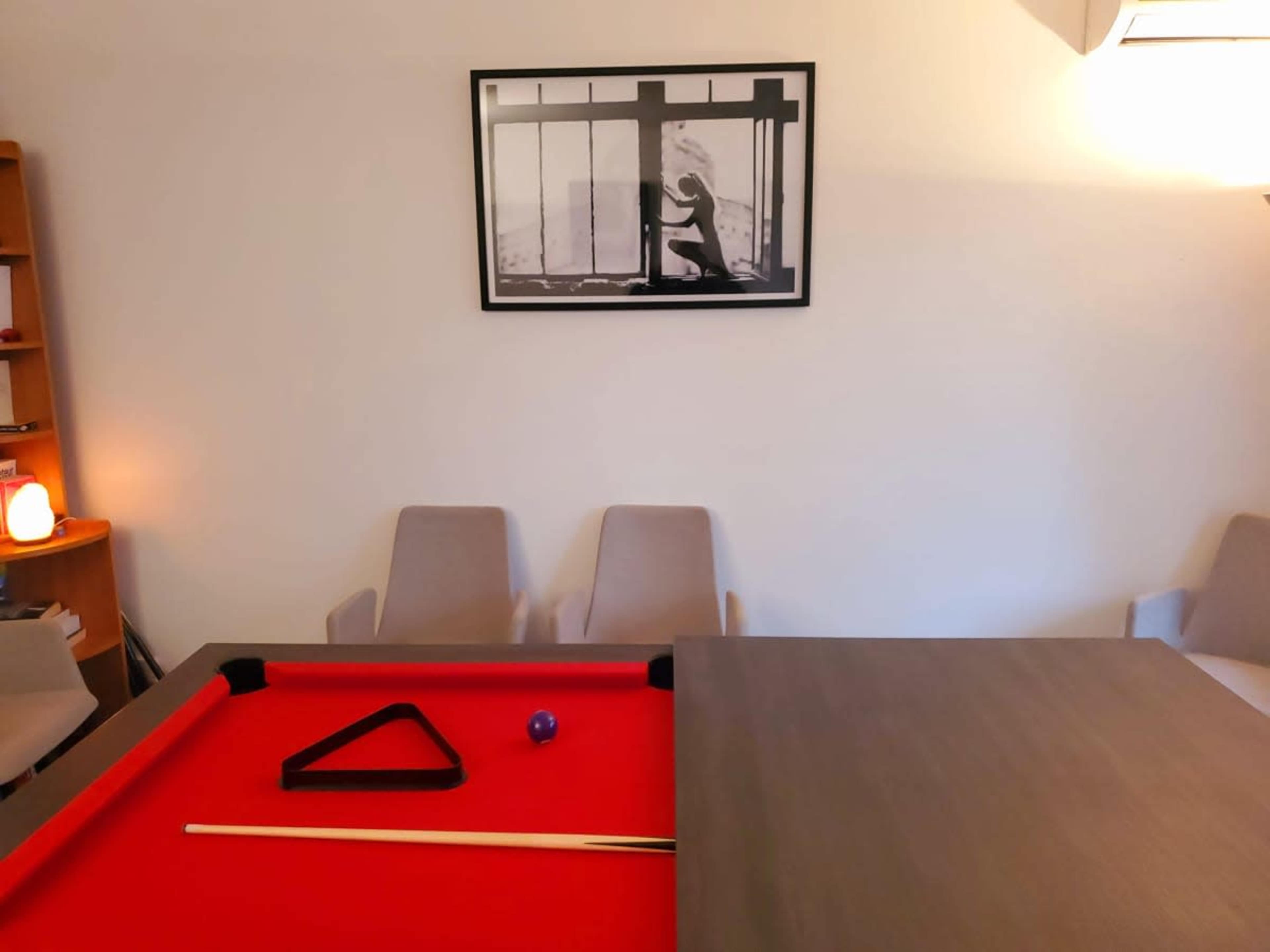 The image shows a pool table with red felt and a triangle rack, positioned in a room with two light-colored chairs and a wall-mounted black-and-white photograph.