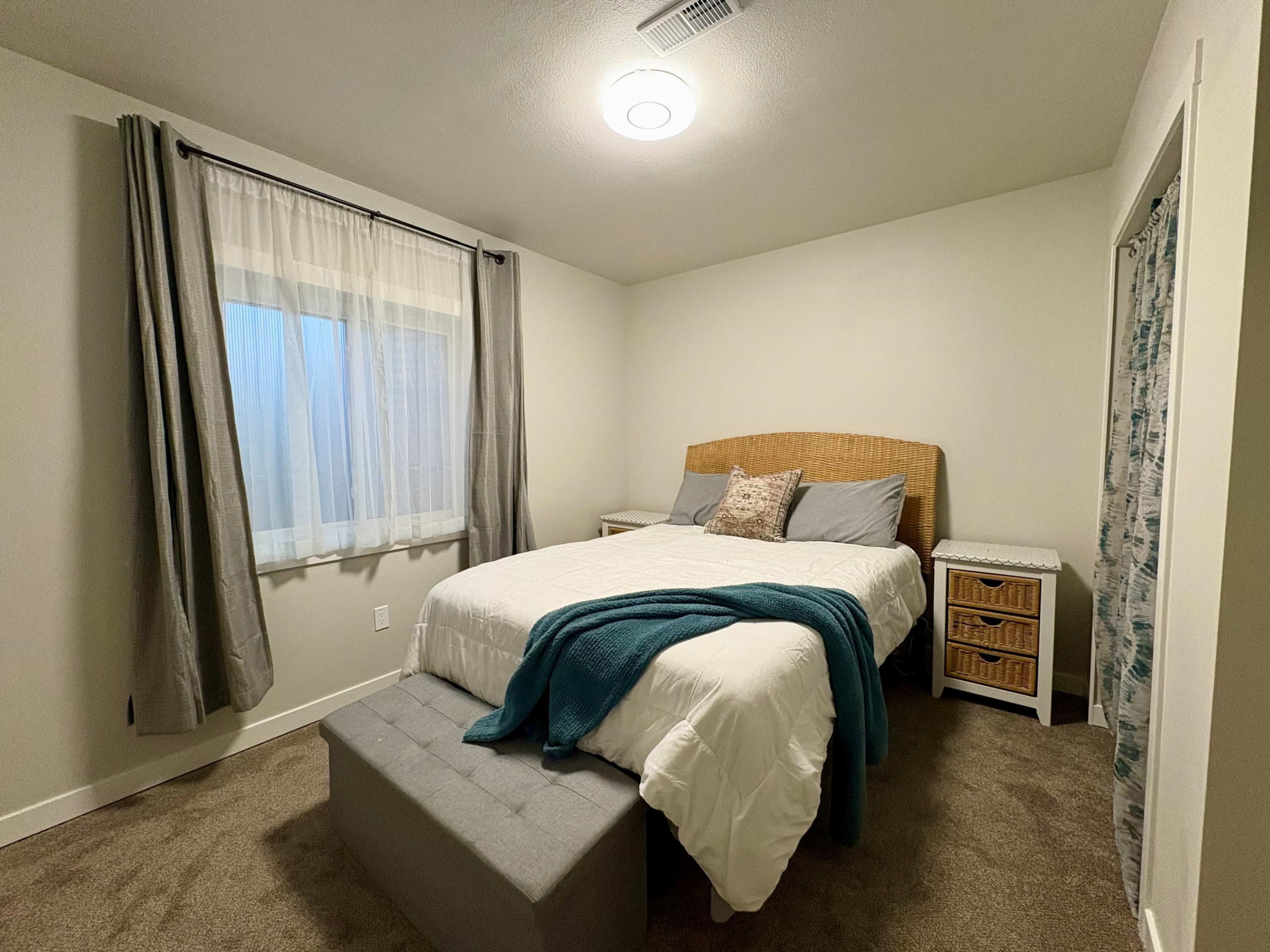 The image shows a simple bedroom with a bed, a nightstand, and a window with sheer curtains.