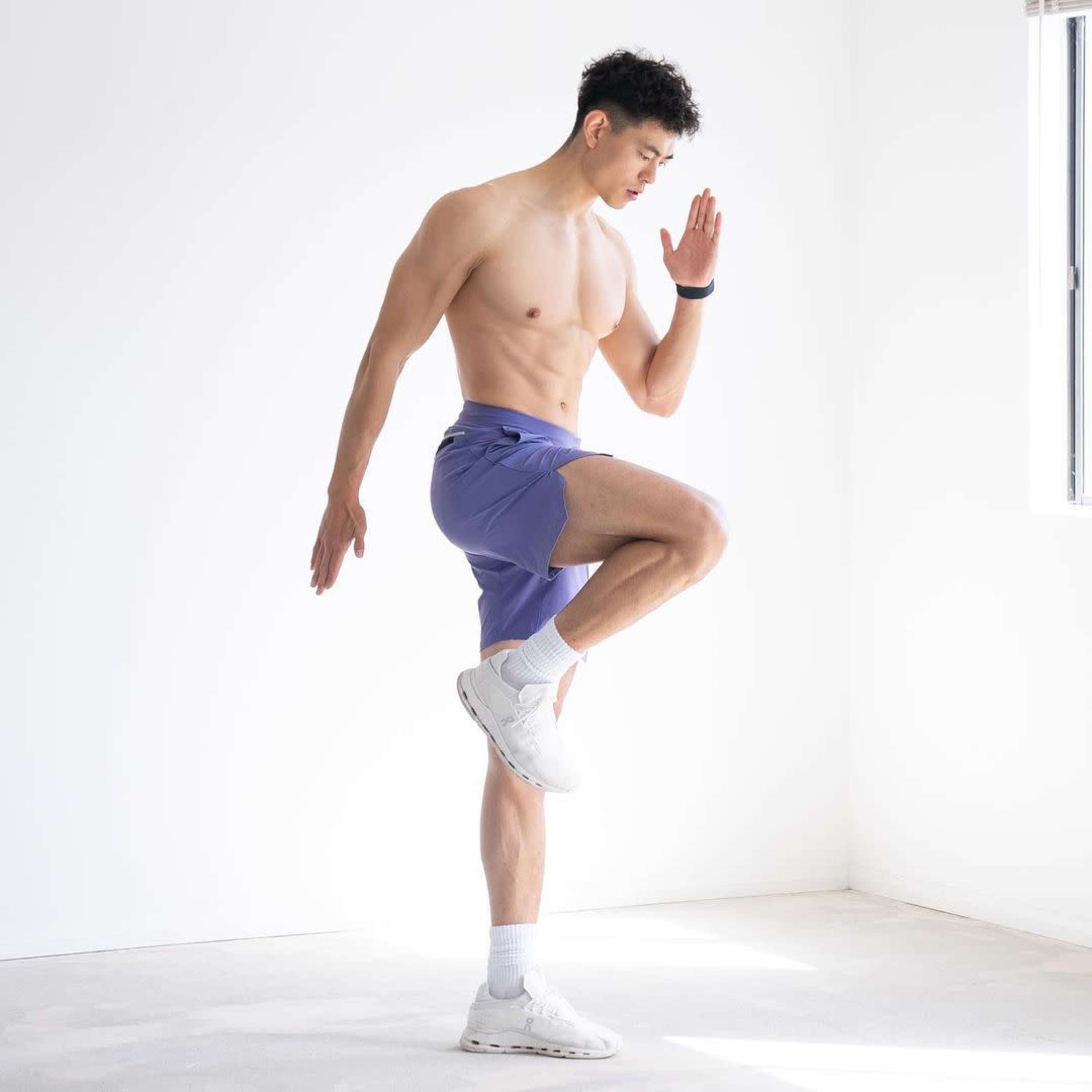 A man in shorts performs a standing exercise, lifting one knee while balancing on the other leg in a bright, minimalist room.