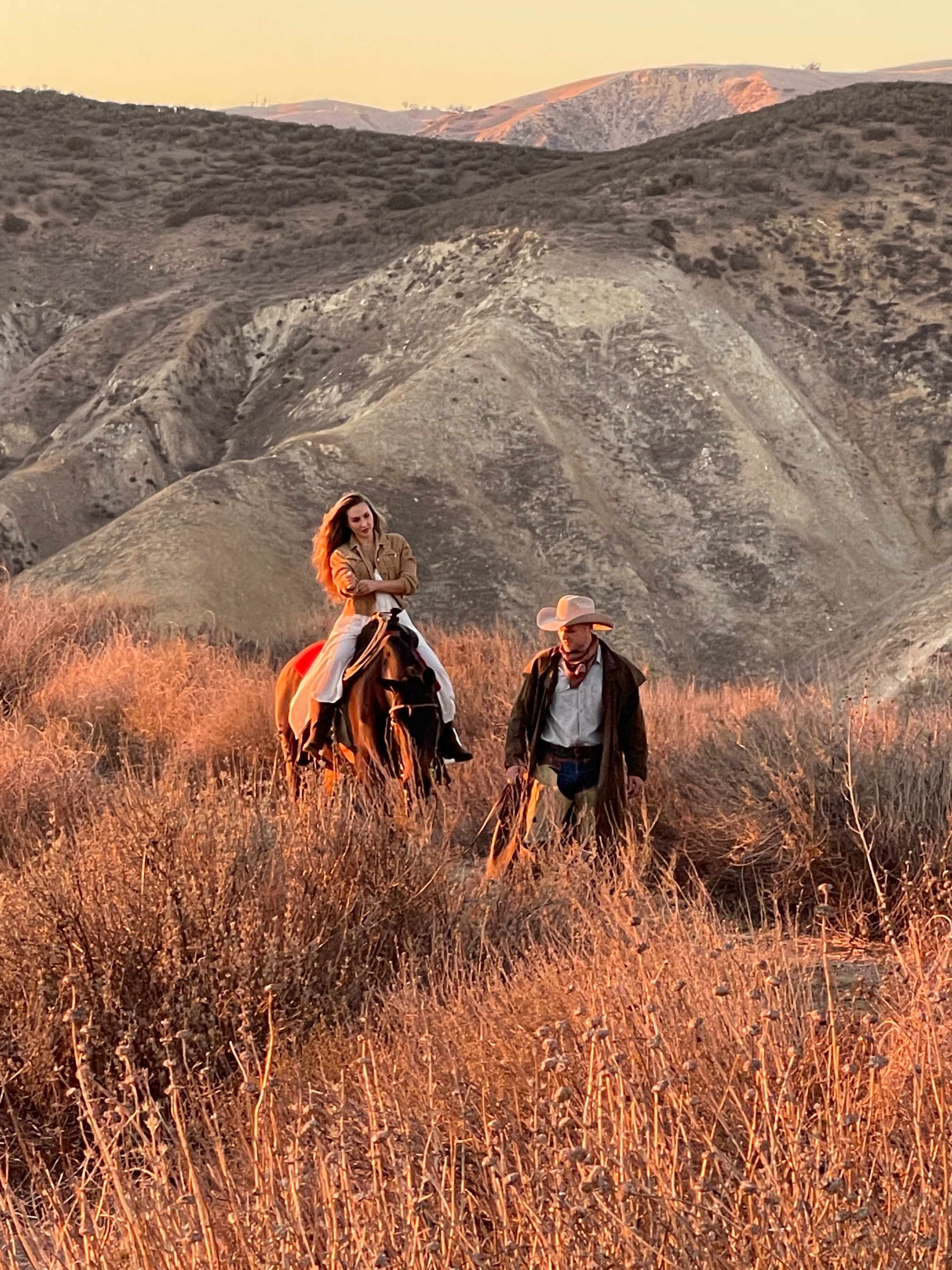 A woman rides a horse while a man walks beside them in a grassy, hilly landscape during sunset.