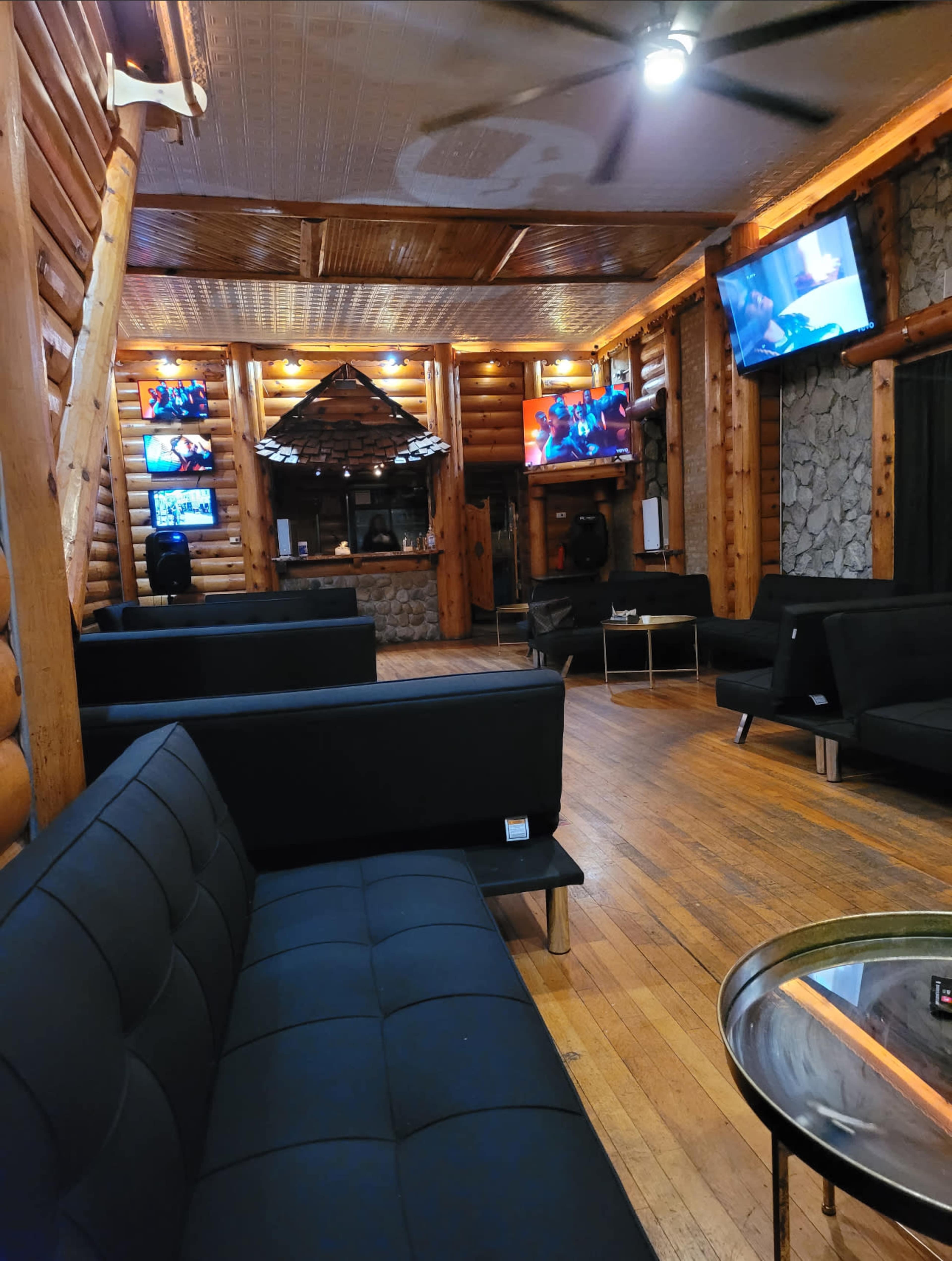 The interior of a log cabin lounge features black sofas and multiple television screens mounted on the walls.