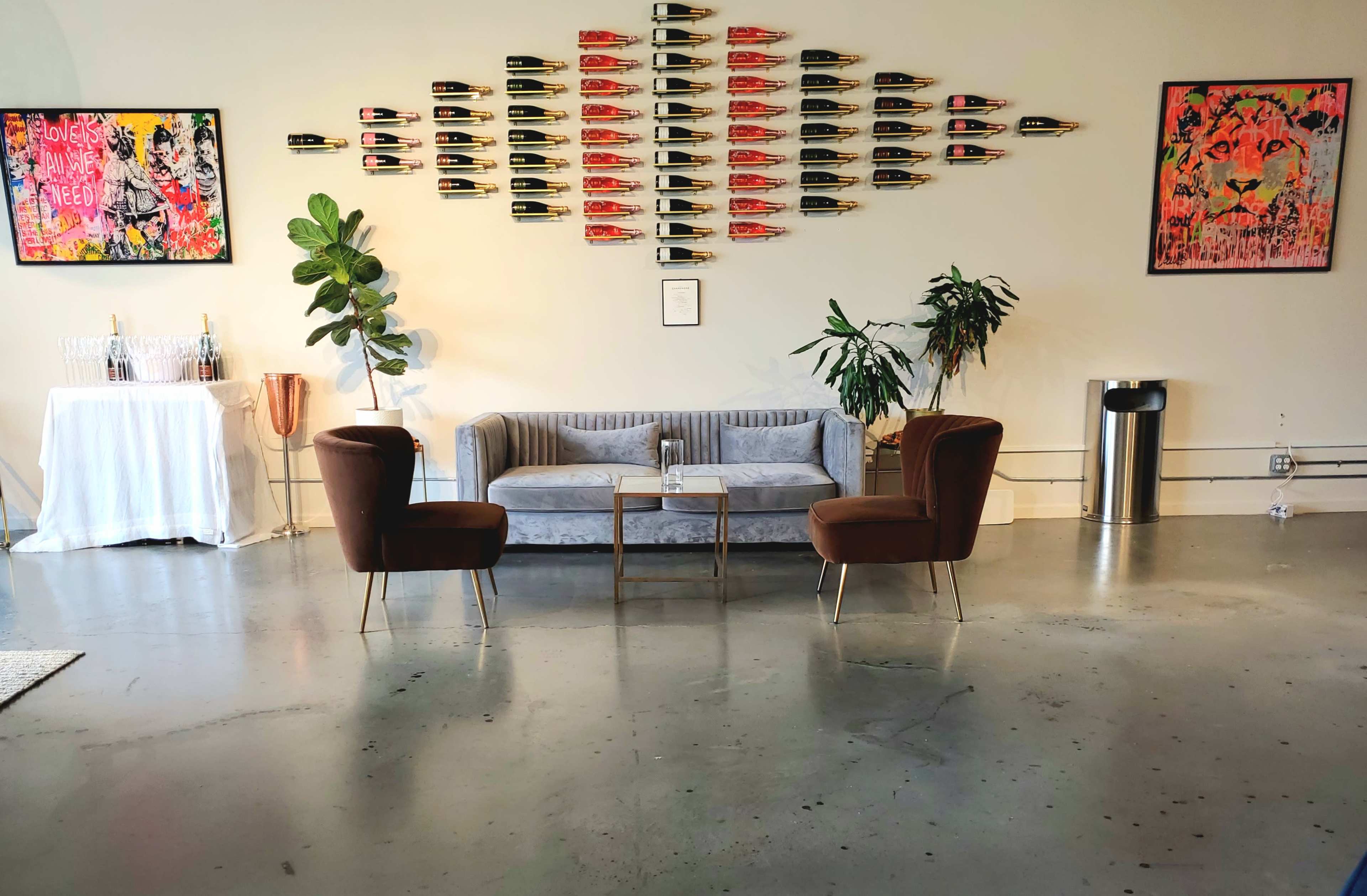 The image shows a modern lounge area with a gray sofa, two brown chairs, a small coffee table, and large art pieces on the walls, including a lion and various graphic artworks.