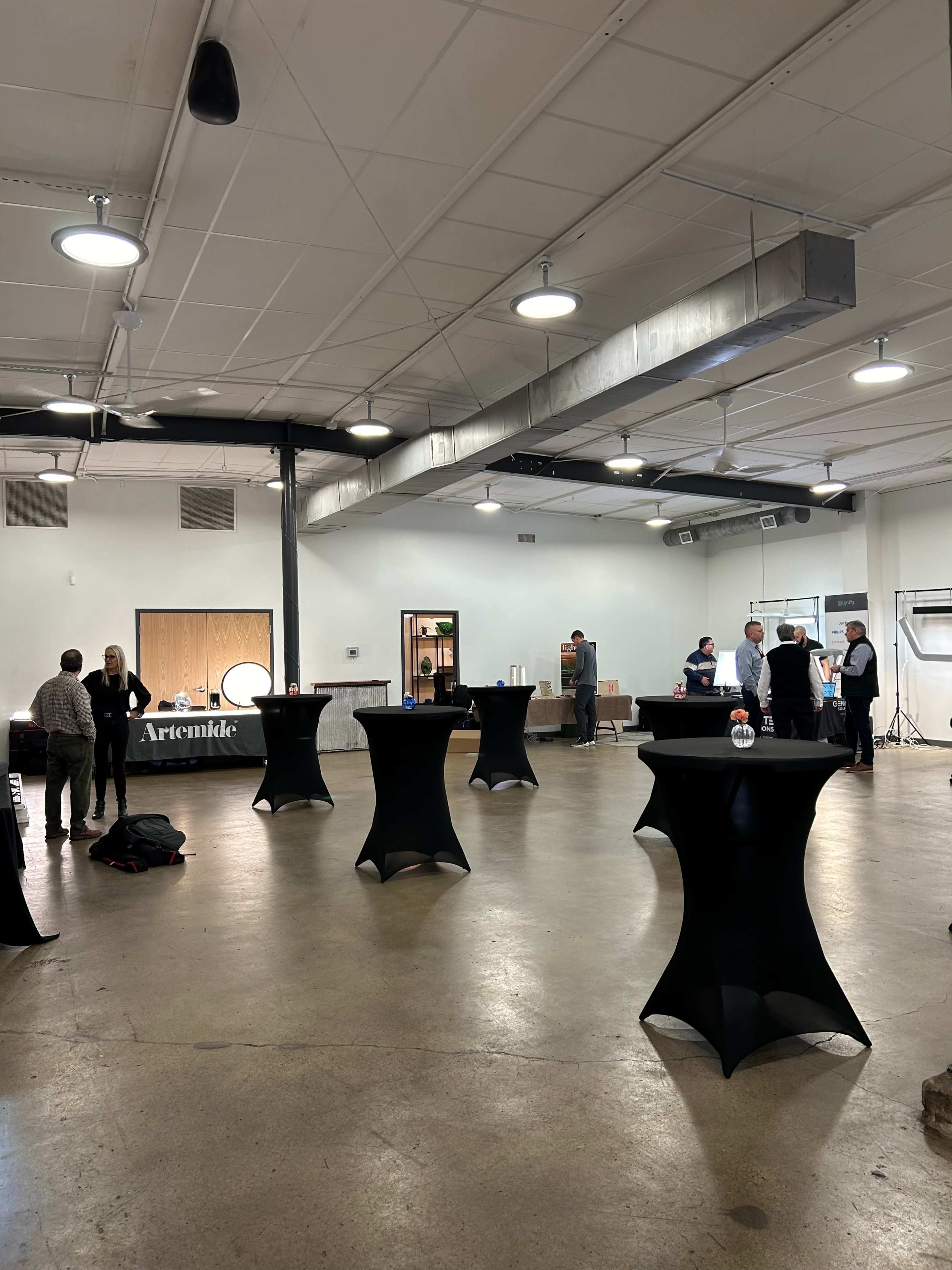 A spacious indoor venue with high ceilings features several black cocktail tables and a small group of people engaged in conversation.