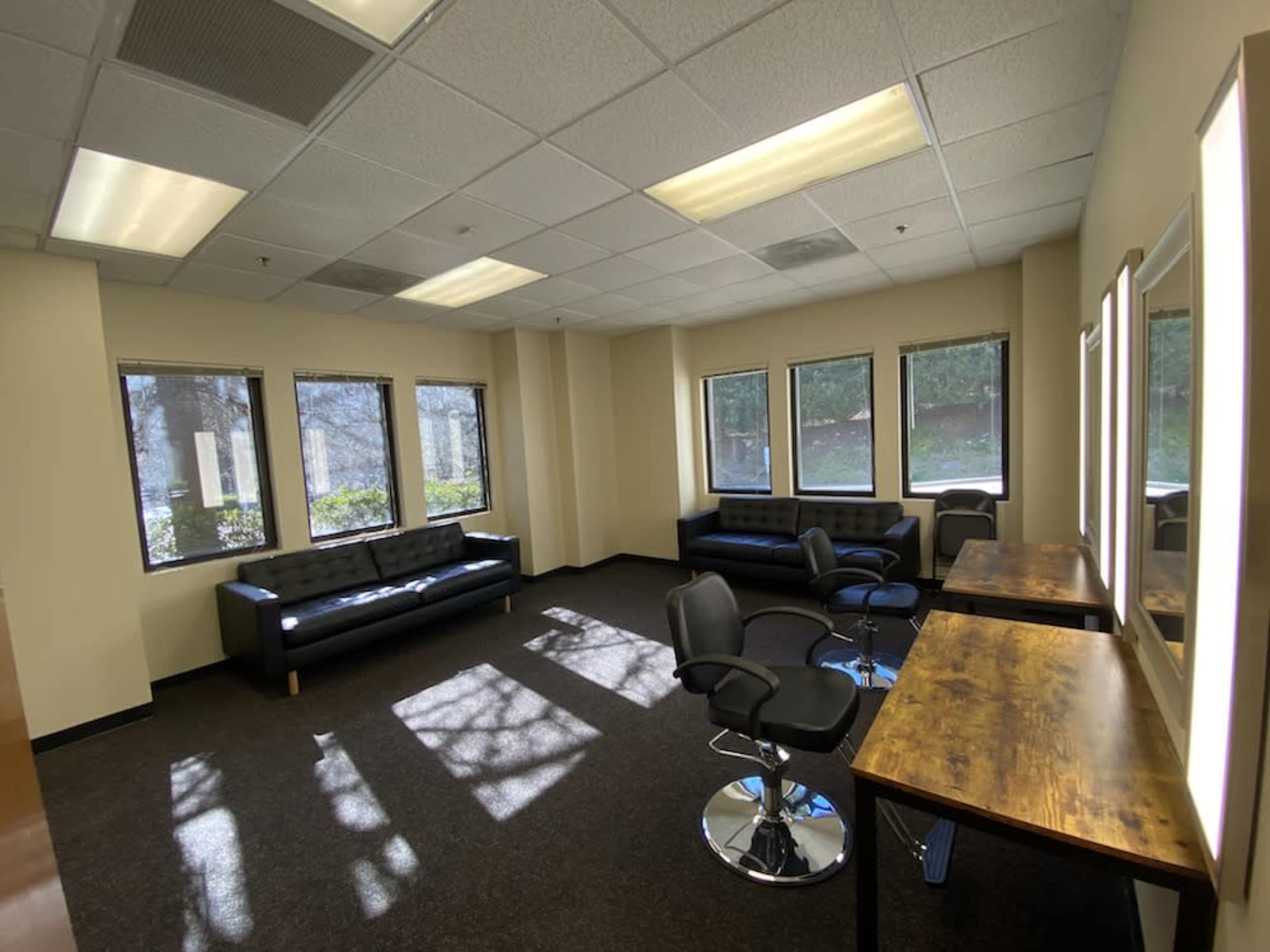 A room with two black sofas, several swivel chairs, and wooden tables, illuminated by natural light from large windows.