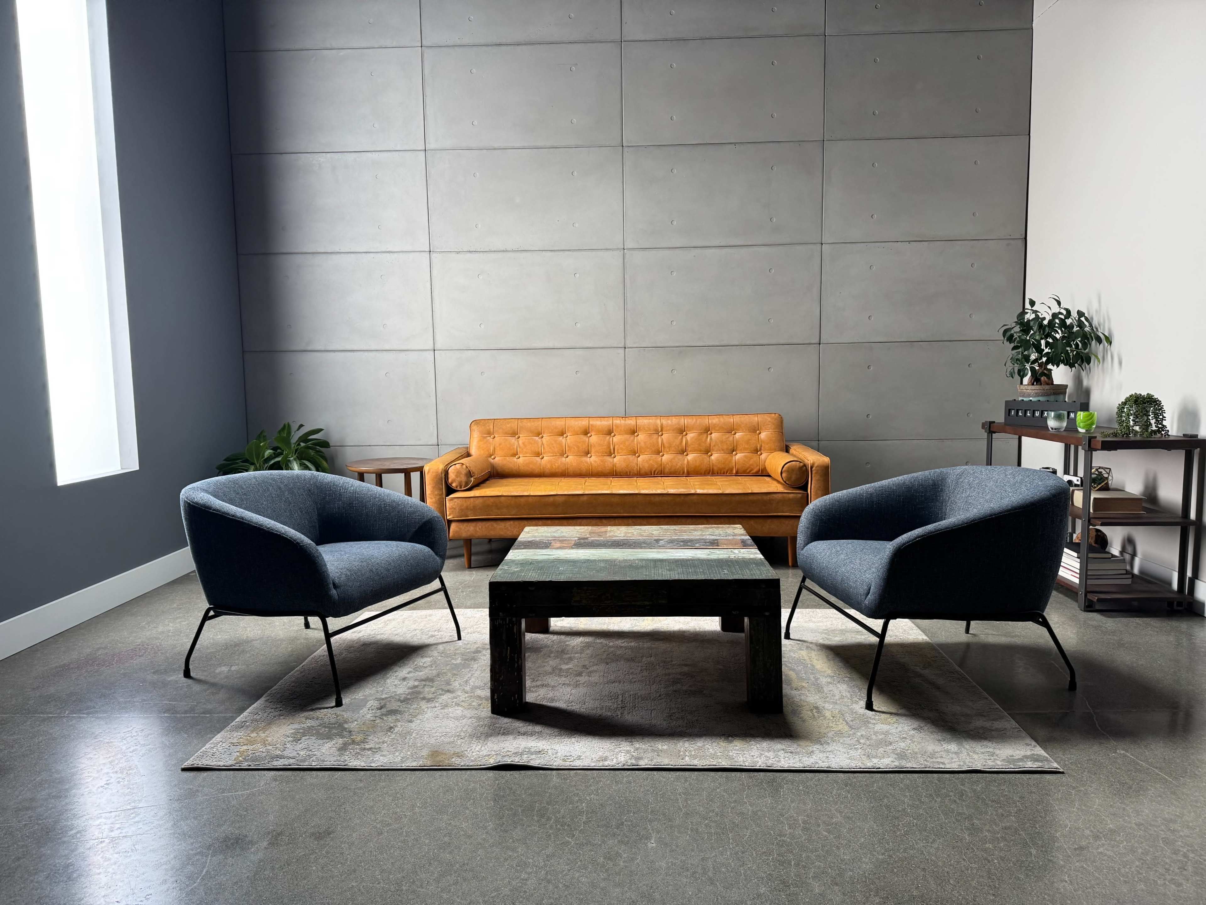 The image shows a modern living space featuring a brown tufted sofa, two blue armchairs, and a rustic wooden coffee table on a patterned rug, with a plant and a shelving unit in the background.
